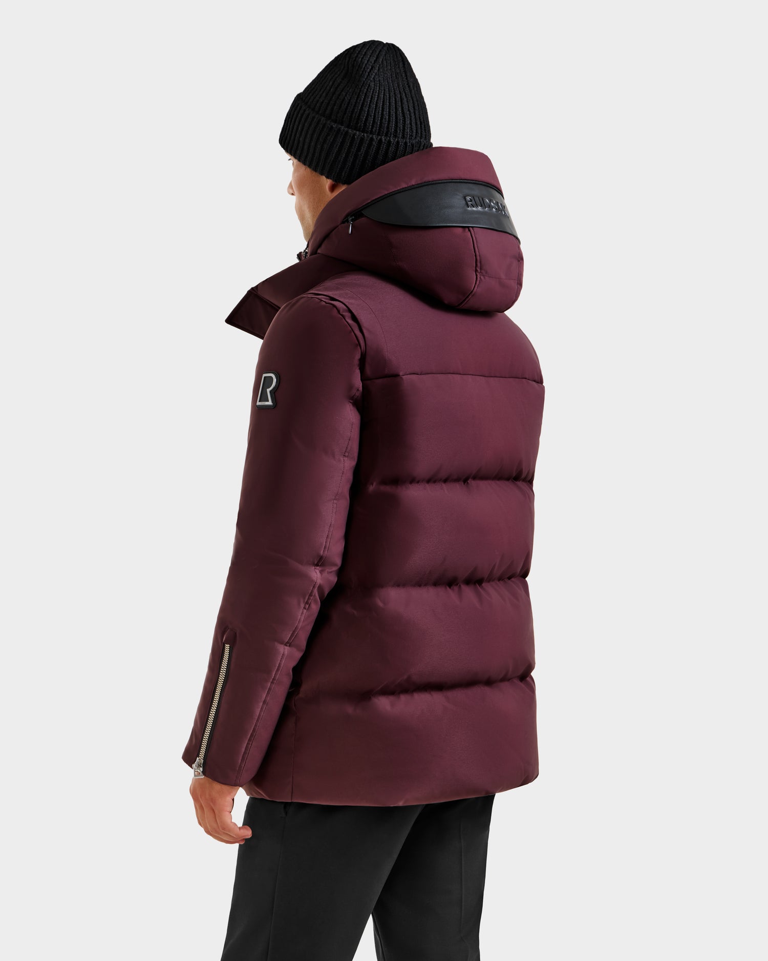 A person wears the Rudsak APOLLO H - 6125690 Bordeaux quilted down parka with a hood, paired with a black beanie and black pants, standing with their back to the camera against a plain light background.