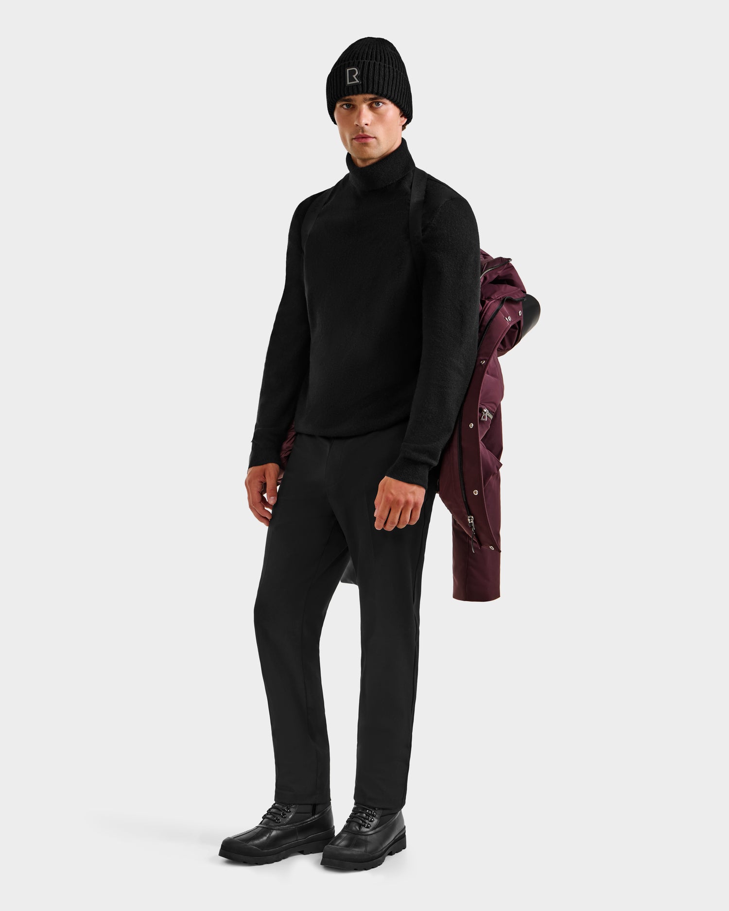 A man in a black turtleneck, pants, shoes, and beanie stands against a white background, holding the Rudsak APOLLO H - 6125690 Bordeaux men’s down parka draped over his shoulder.