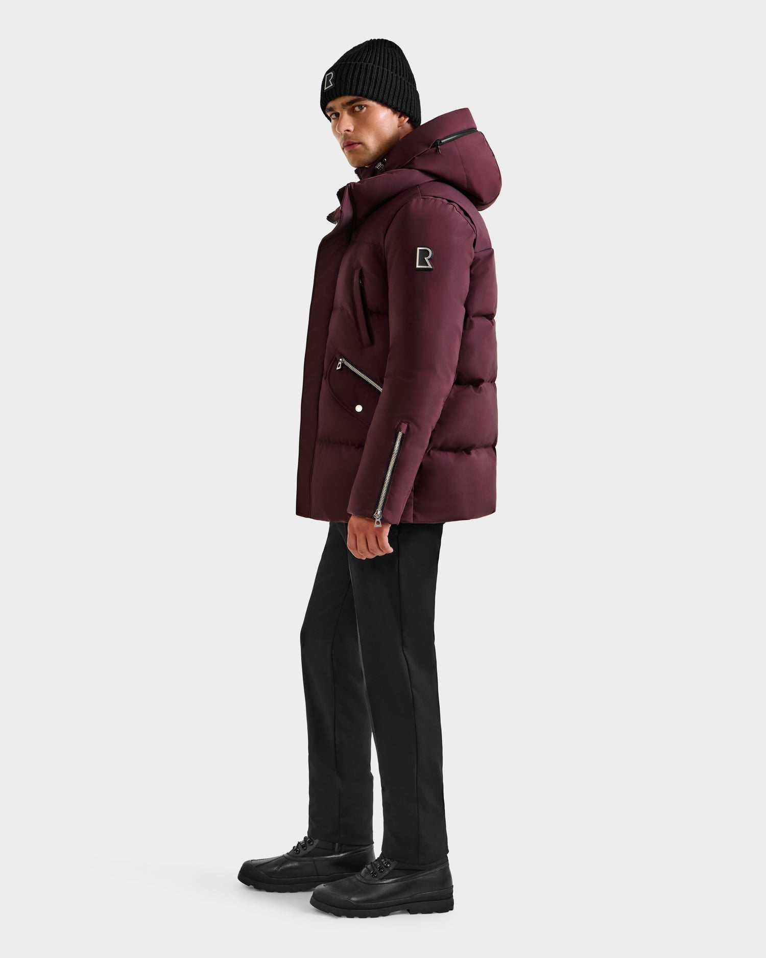 A person wears the Rudsak APOLLO H - 6125690 Bordeaux men’s quilted down parka, black pants, black shoes, and a black beanie while facing left against a plain white background.