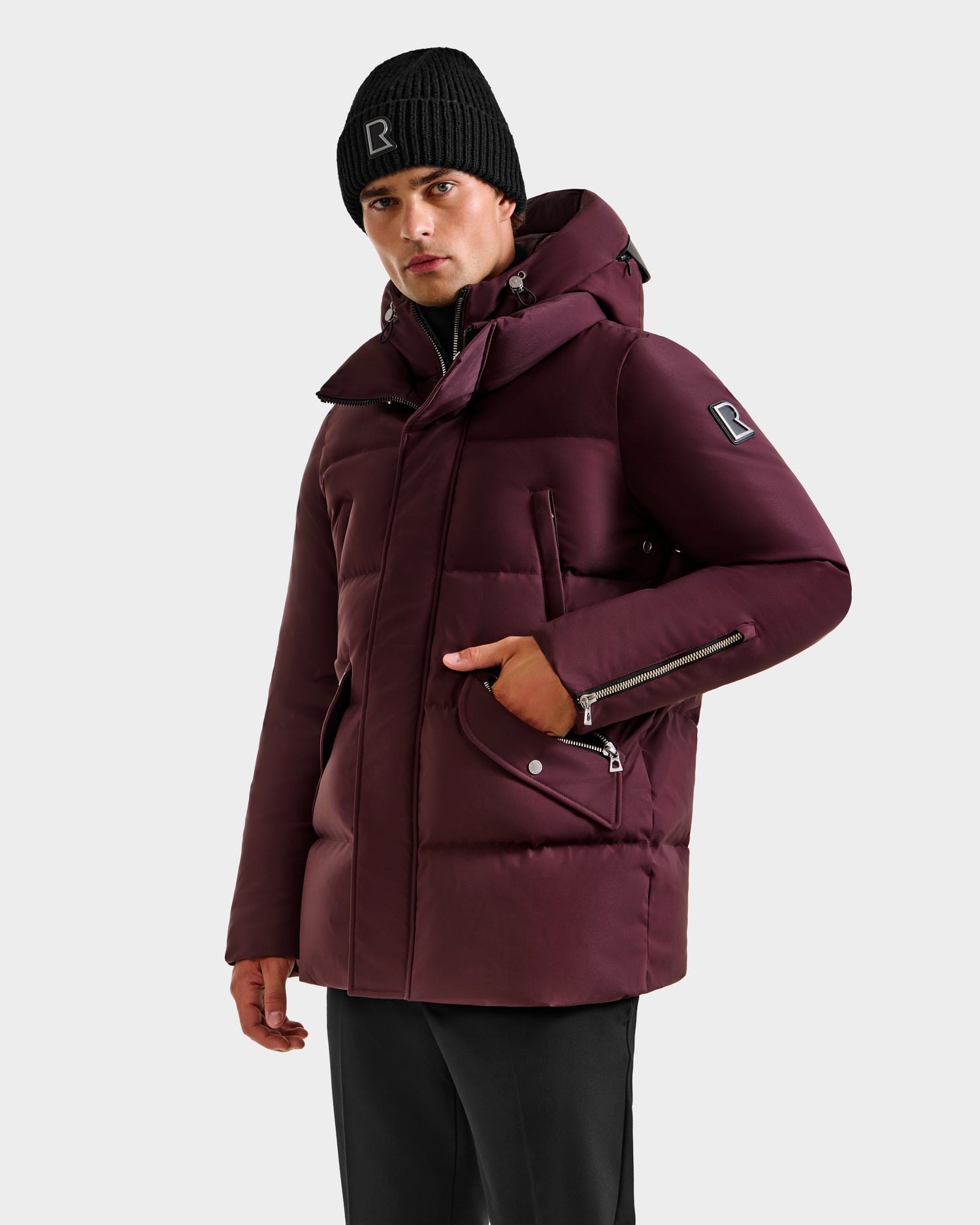 A person wears the Rudsak APOLLO H - 6125690 Bordeaux men's parka with hood and zippered pockets, paired with a black beanie and pants, standing against a plain light background.