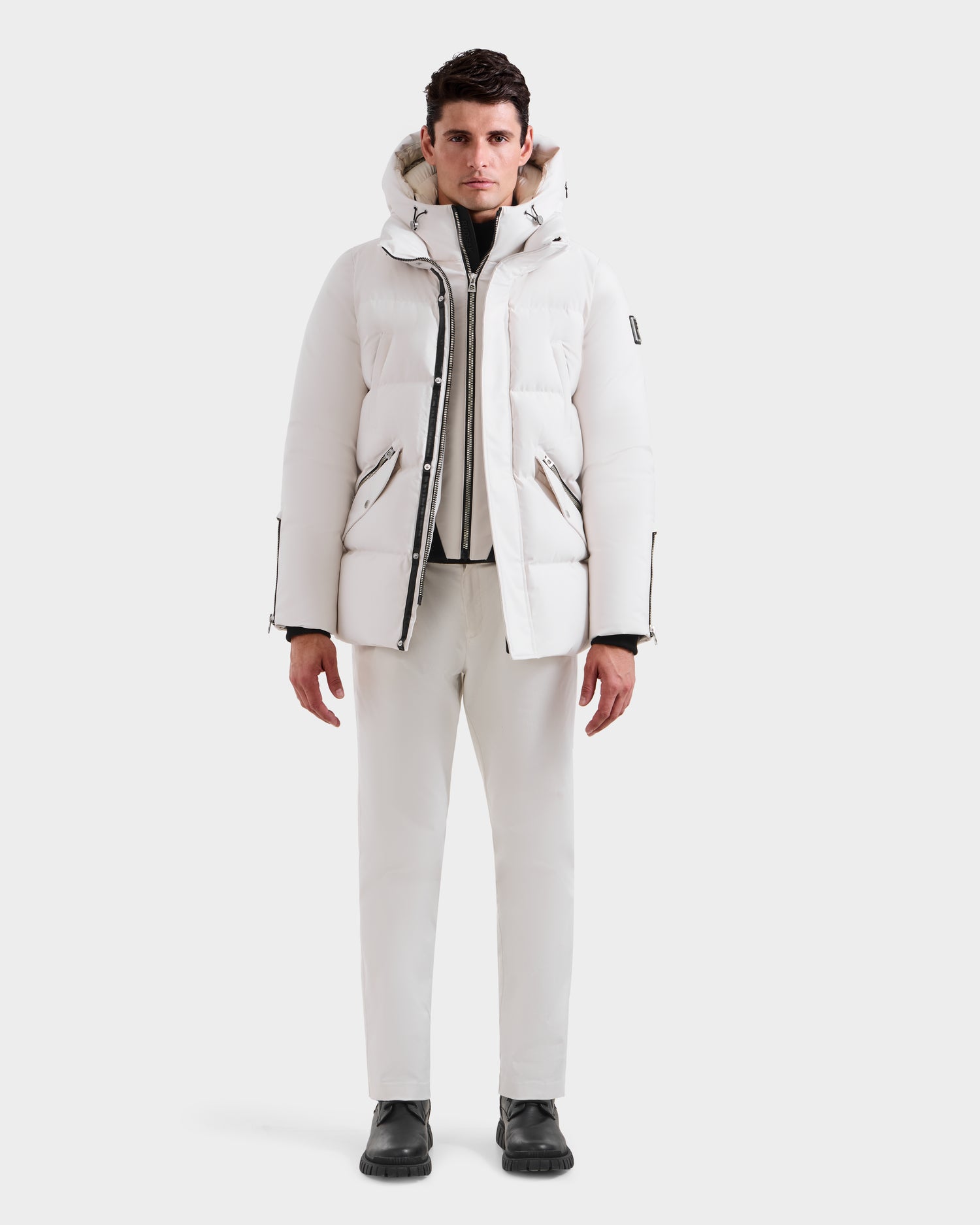 A man stands facing forward in an all-white winter outfit, featuring the Rudsak APOLLO H - 6125690 Vanilla men’s down parka with a hood, matching pants, and black boots, set against a plain white background.