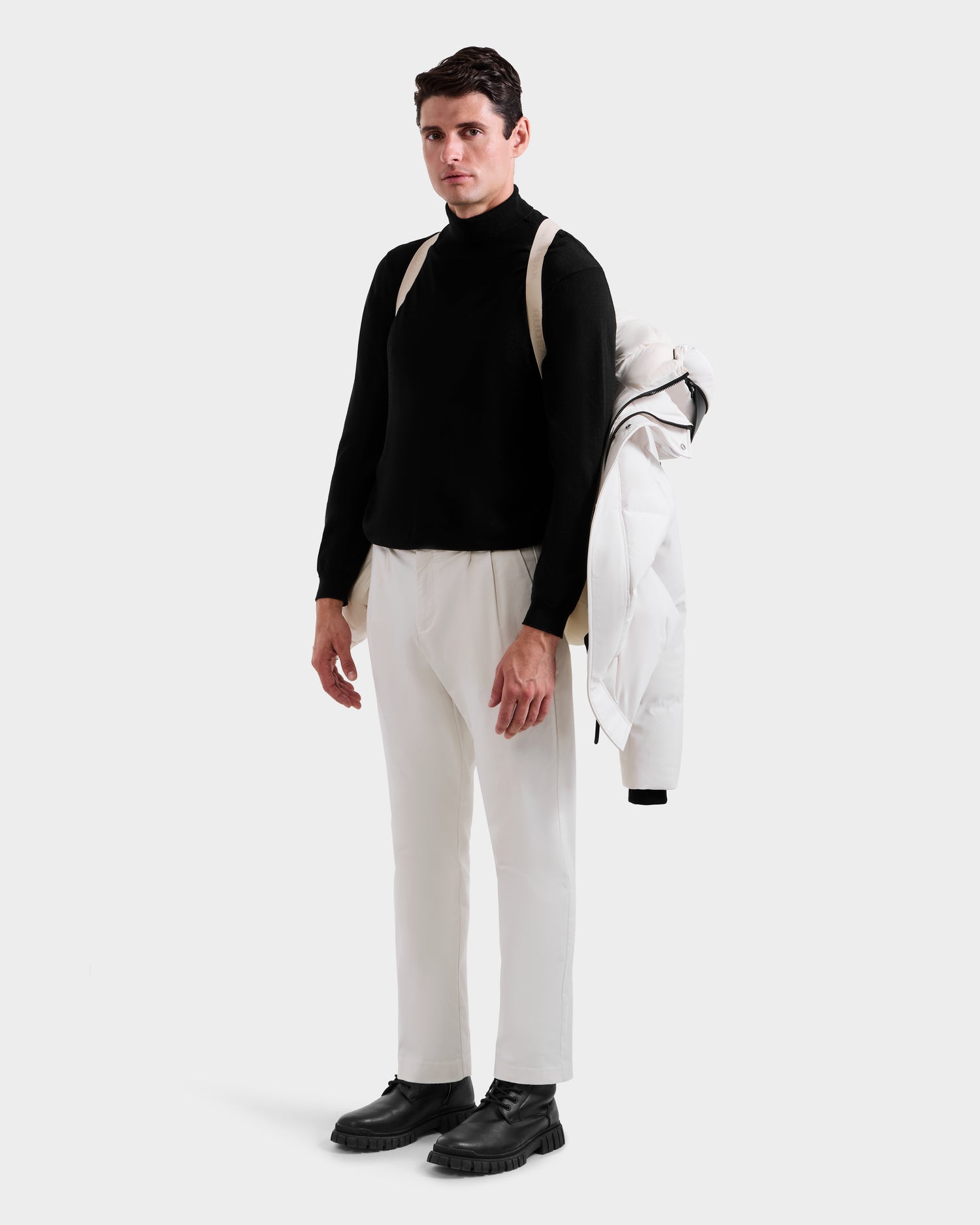 A man stands on a white background, wearing a black turtleneck, white pants, black boots, and holding the Rudsak APOLLO H - 6125690 Vanilla winter performance parka over his left shoulder while carrying a beige backpack.