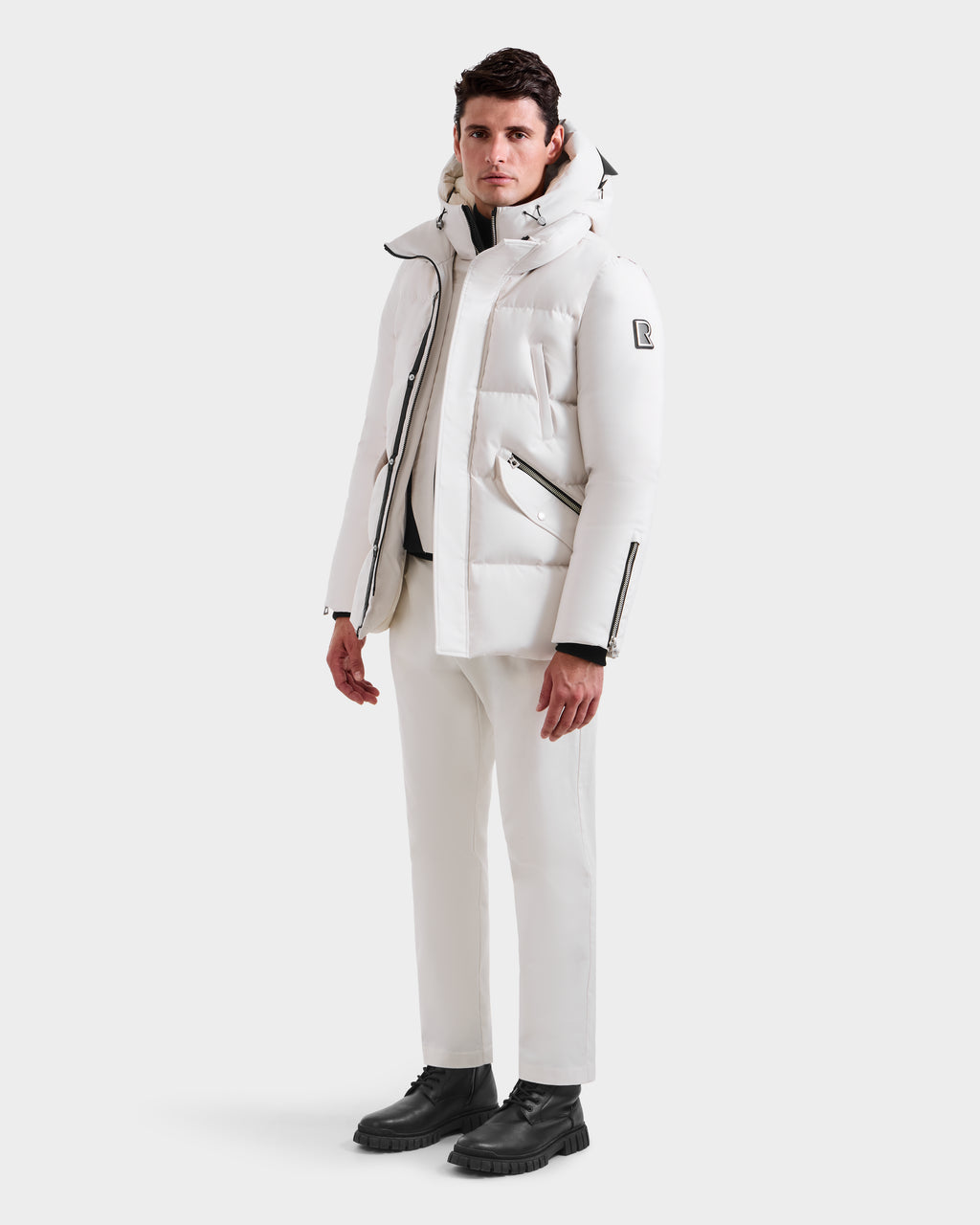 A man faces forward against a plain white background, wearing the Rudsak APOLLO H - 6125690 Vanilla men’s quilted down parka, paired with white pants and black boots.