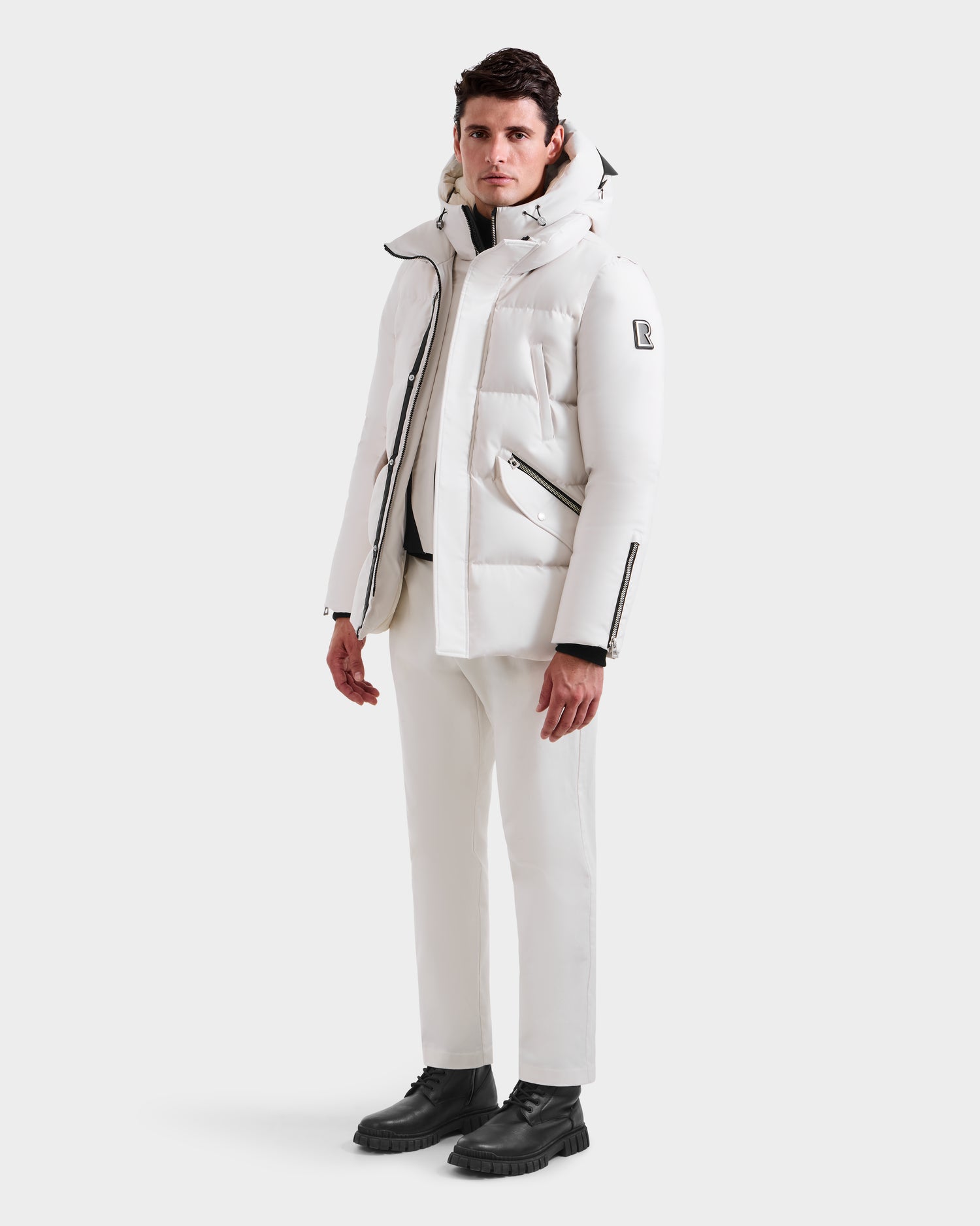 A man faces forward against a plain white background, wearing the Rudsak APOLLO H - 6125690 Vanilla men’s quilted down parka, paired with white pants and black boots.