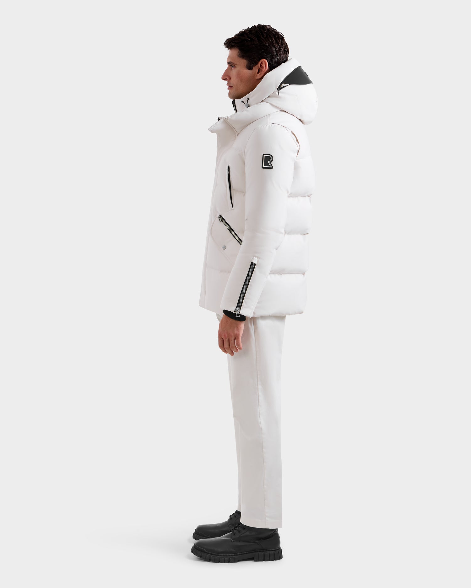 A man in profile against a white background wears the Rudsak APOLLO H - 6125690 Vanilla quilted parka, featuring a hood, black B patch on the sleeve, multiple zippered pockets, white pants, and black boots.