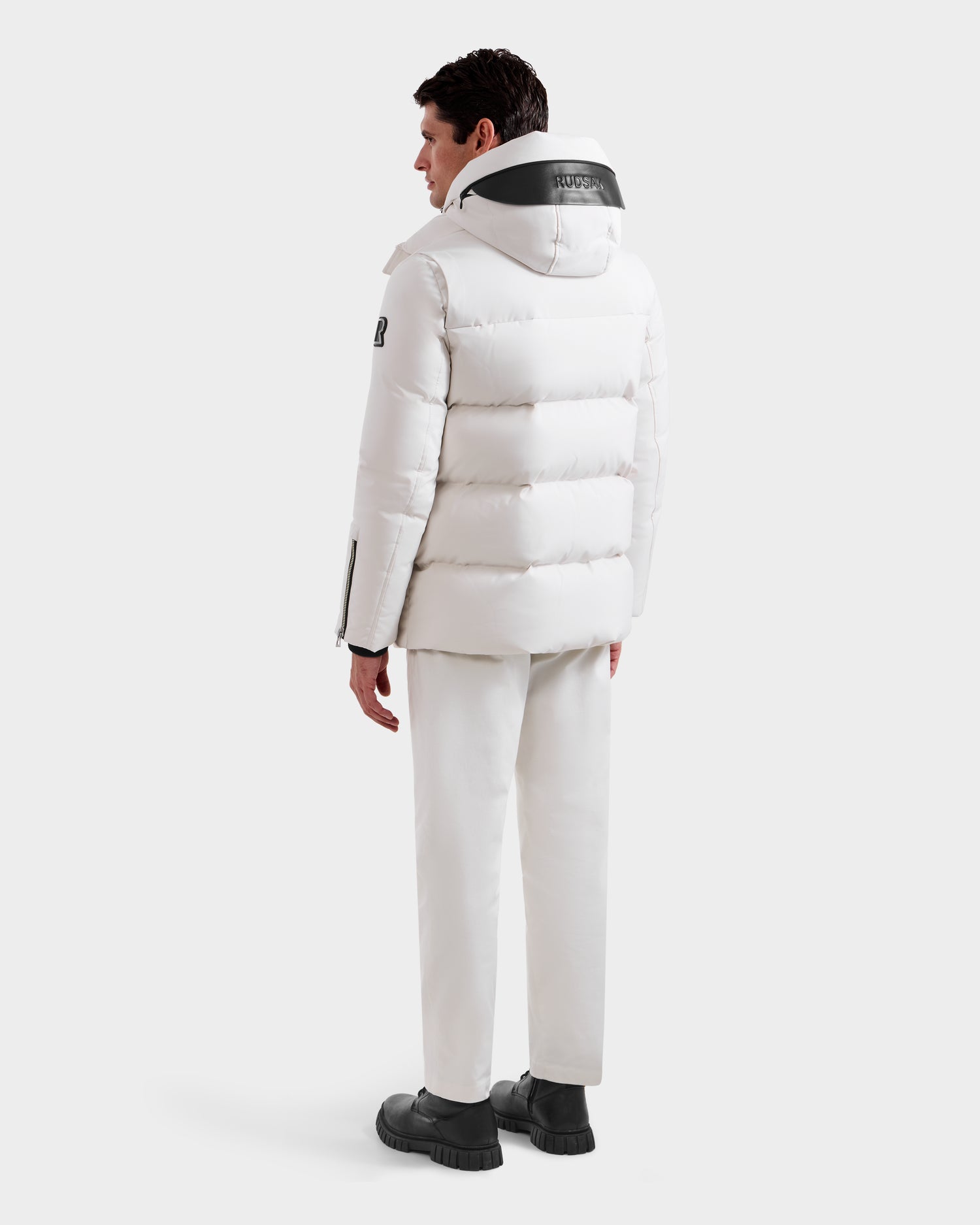 A person stands facing away, wearing the Rudsak APOLLO H - 6125690 Vanilla men’s quilted down parka with vegan leather trims, styled with white pants and black shoes against a plain light background.