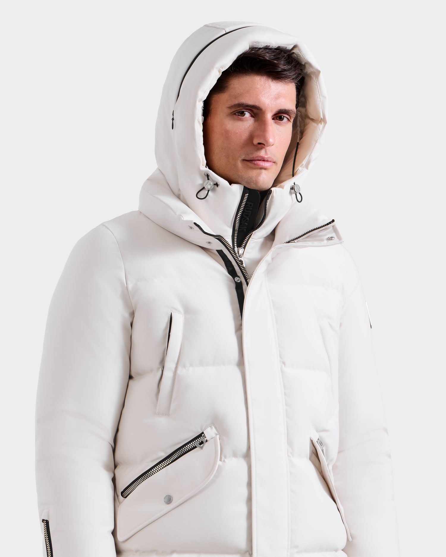 A man wears the Rudsak APOLLO H - 6125690 Vanilla, a white quilted winter performance parka with hood up, zipper pockets, and high collar, looking to the side against a plain white background.