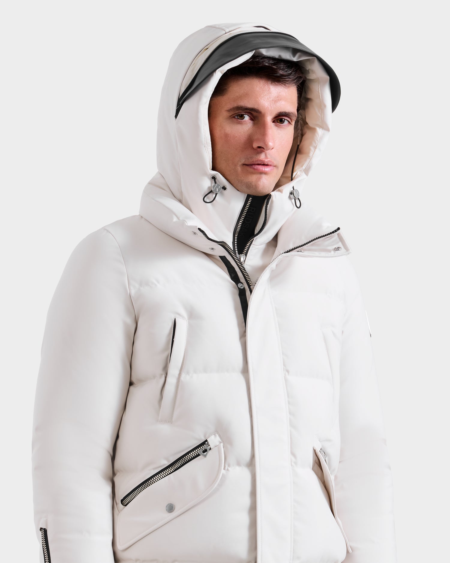 A man stands against a plain light background wearing the Rudsak APOLLO H - 6125690 Vanilla men’s quilted down parka, featuring black zipper details and a partially zipped front.