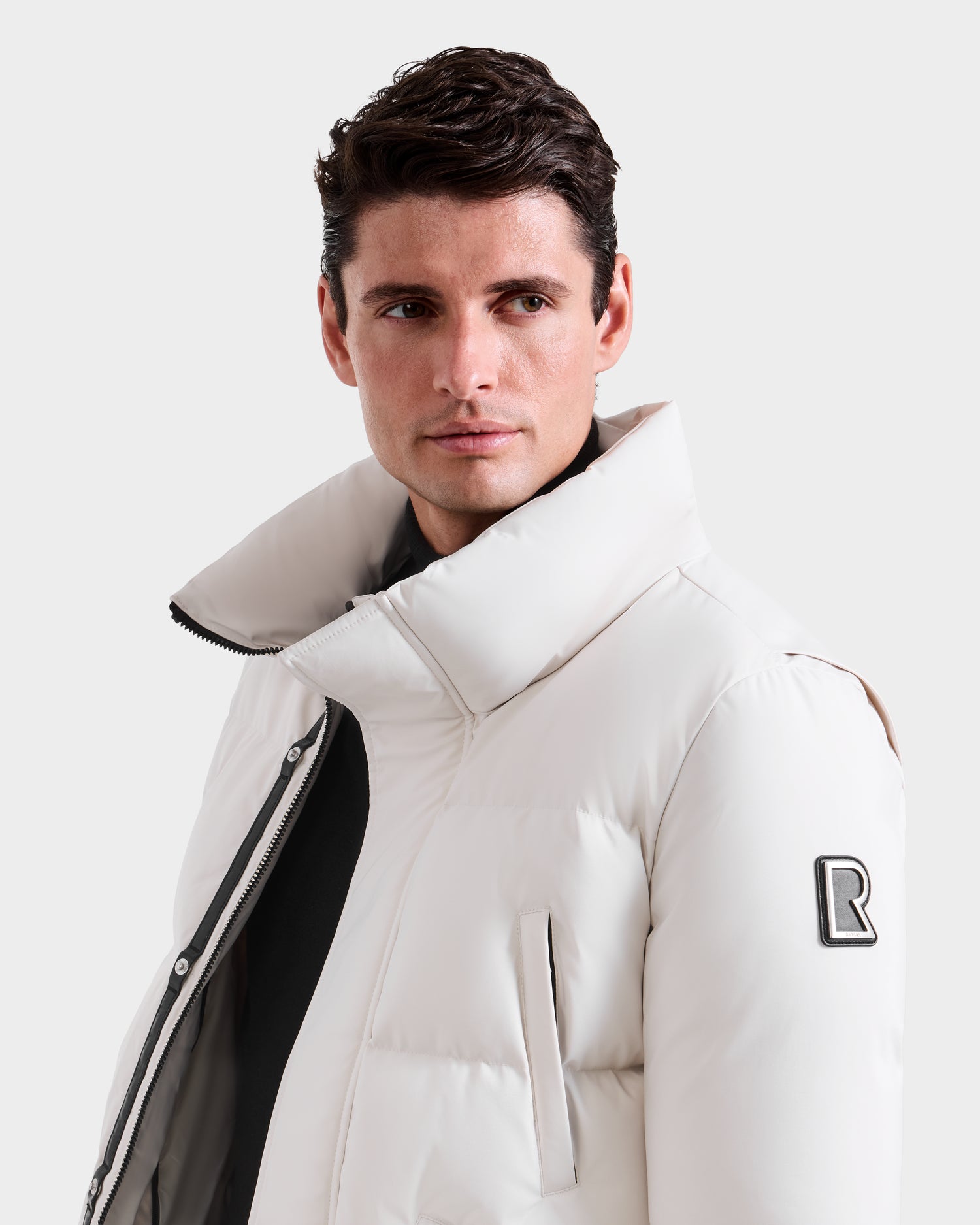 A man with short brown hair wears the Rudsak APOLLO H - 6125690 Vanilla winter jacket, featuring a high collar and black R patch on the sleeve, looking slightly left against a plain light background.