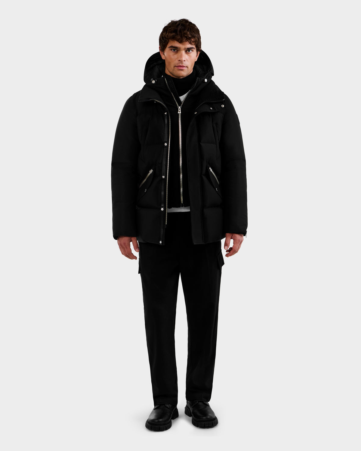 A man stands facing forward, wearing the Rudsak APOLLO - 6125693 Black hooded puffer jacket over a black turtleneck, paired with black trousers and shoes, set against a plain light gray background.