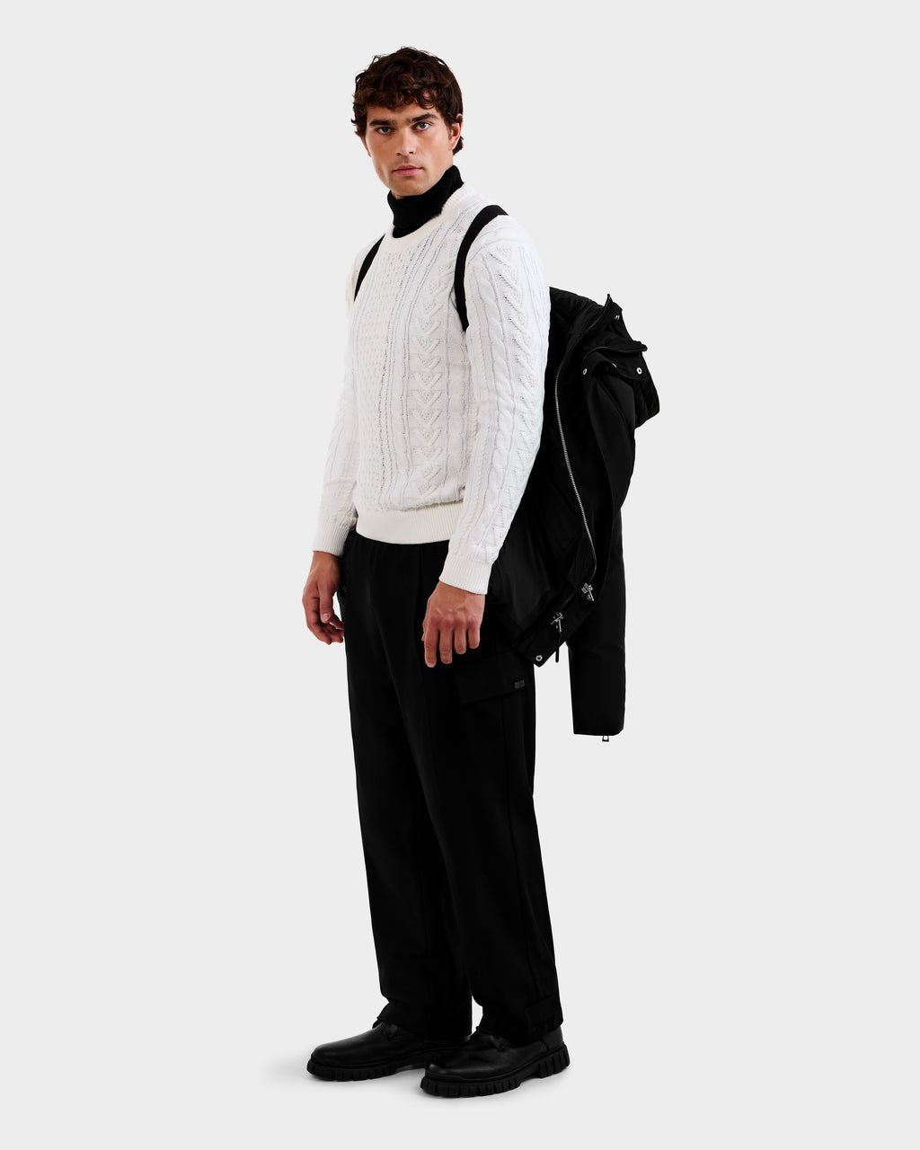 A man stands against a plain light background, holding the Rudsak APOLLO - 6125693 Black jacket over his shoulder, dressed in a white cable-knit sweater, black pants, and black shoes, gazing slightly to his right.