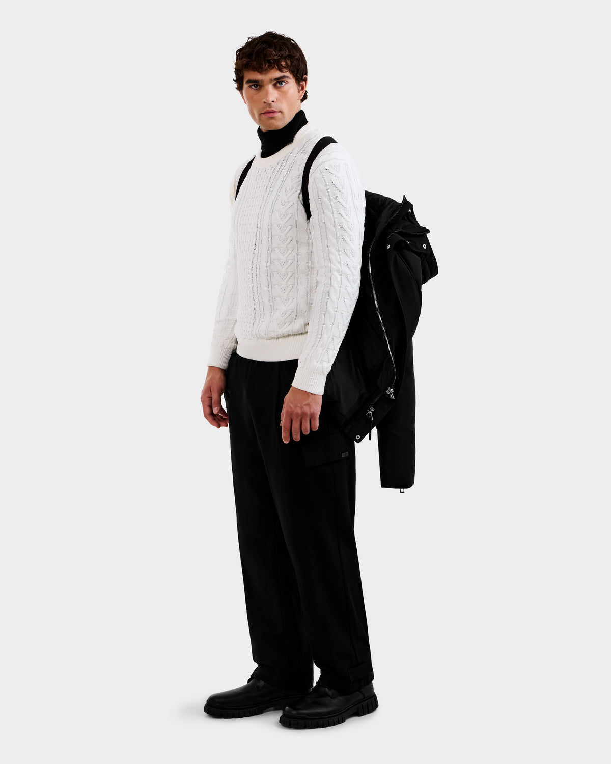 A man stands against a plain light background, holding the Rudsak APOLLO - 6125693 Black jacket over his shoulder, dressed in a white cable-knit sweater, black pants, and black shoes, gazing slightly to his right.