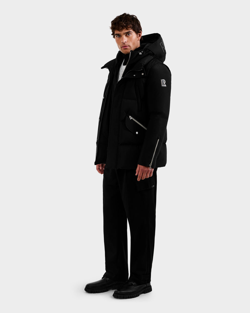 A man stands slightly angled, wearing the Rudsak APOLLO - 6125693 Black hooded winter jacket, paired with black pants and shoes against a plain white background.