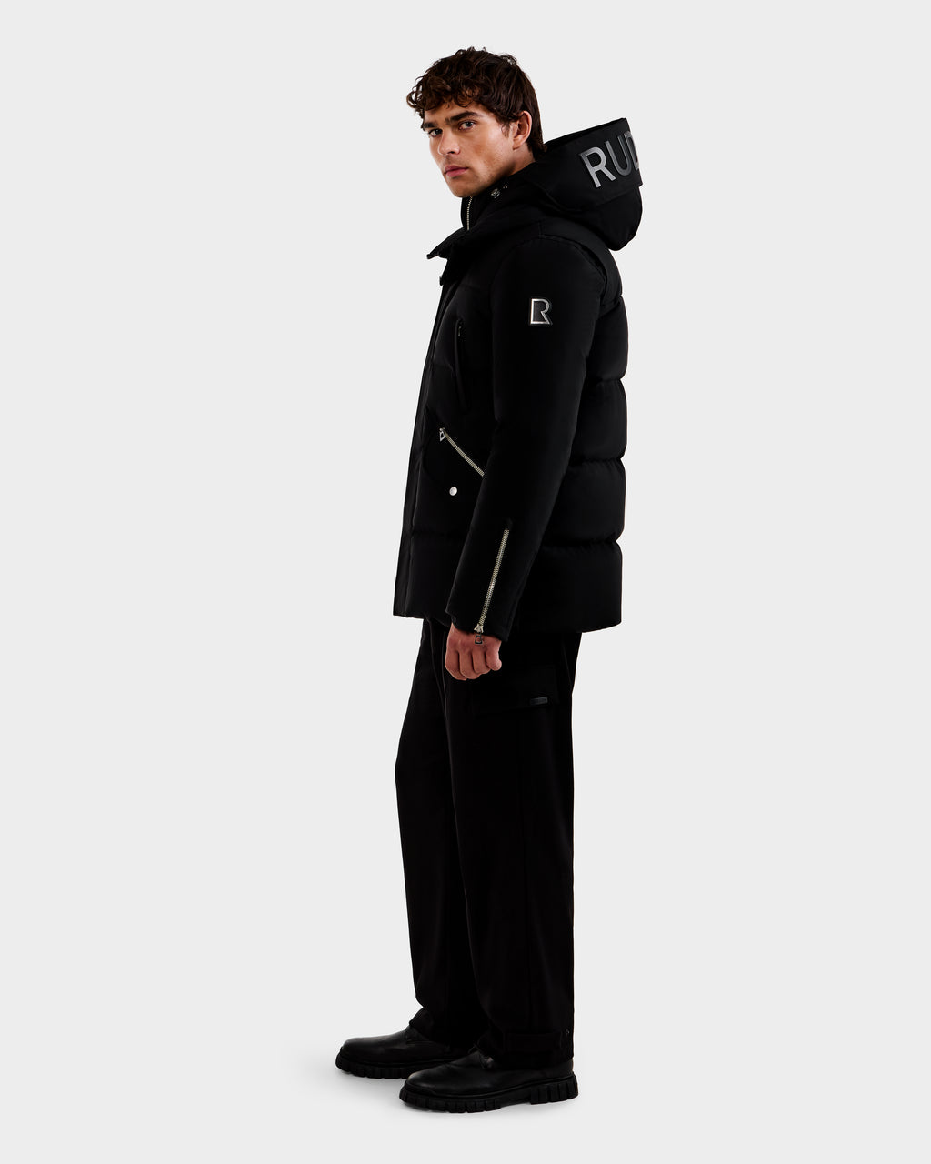 A man stands sideways against a white background, wearing the Rudsak APOLLO - 6125693 Black puffer jacket with a hood featuring white lettering, paired with black pants and black boots.