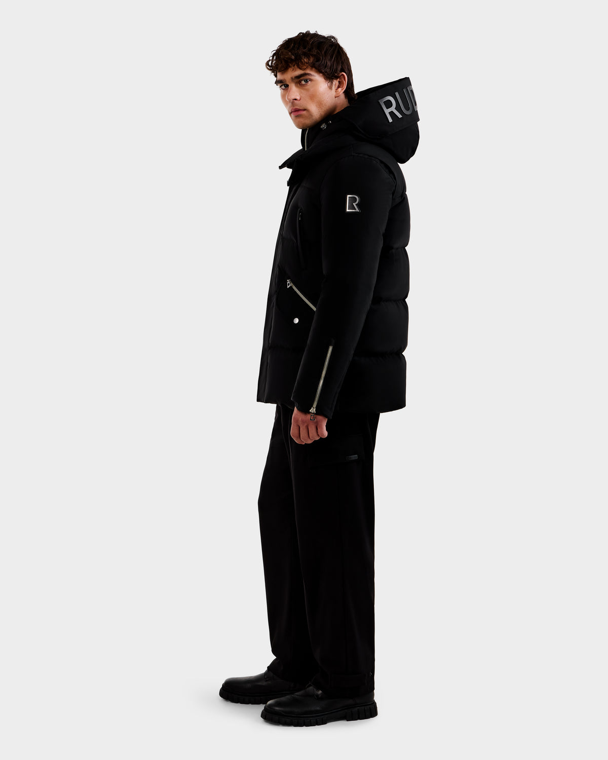 A man stands sideways against a white background, wearing the Rudsak APOLLO - 6125693 Black puffer jacket with a hood featuring white lettering, paired with black pants and black boots.