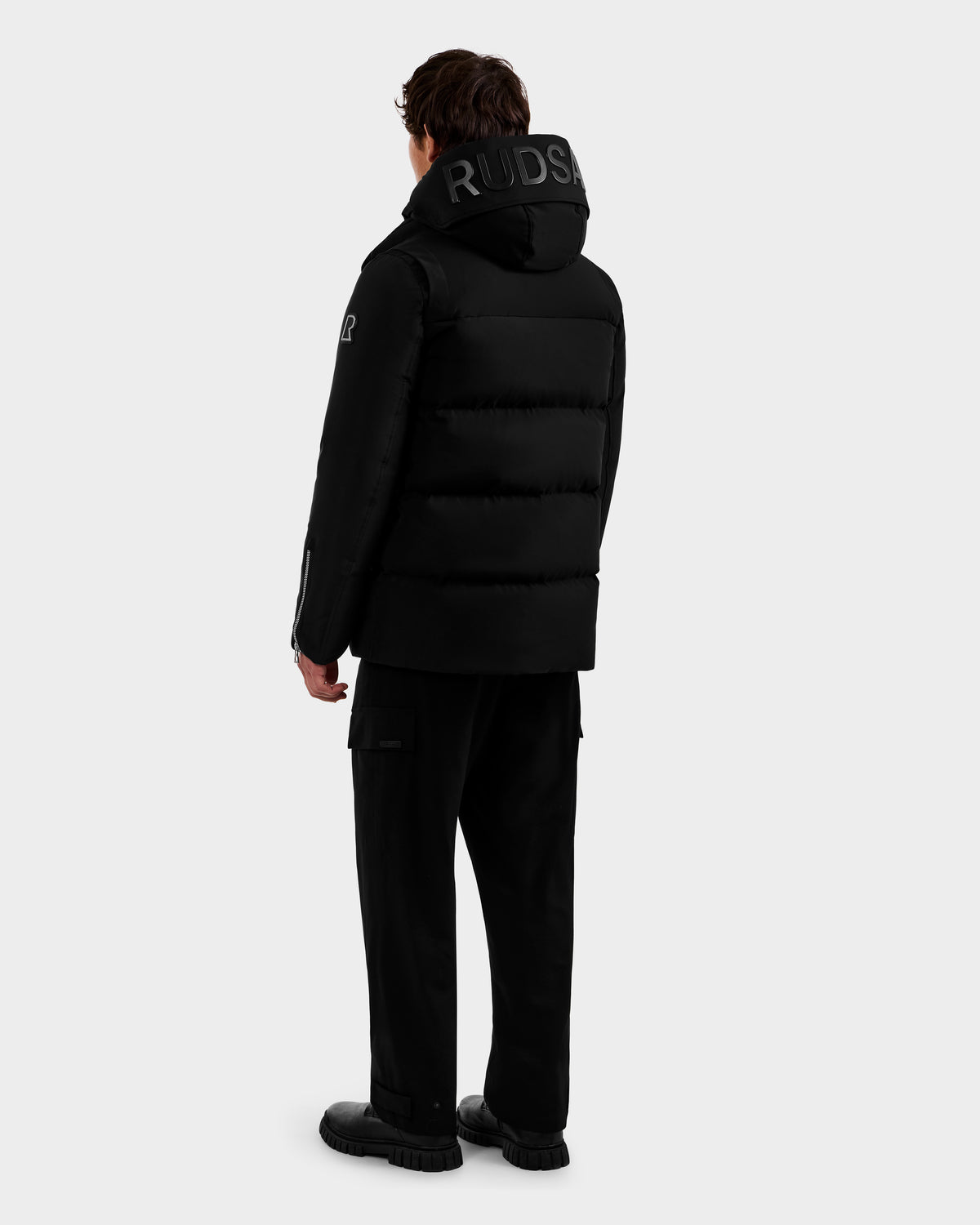 A person stands facing away, wearing the Rudsak APOLLO - 6125693 Black hooded puffer jacket with "RUDSAK" on the hood, paired with black pants and shoes, set against a plain light gray background.