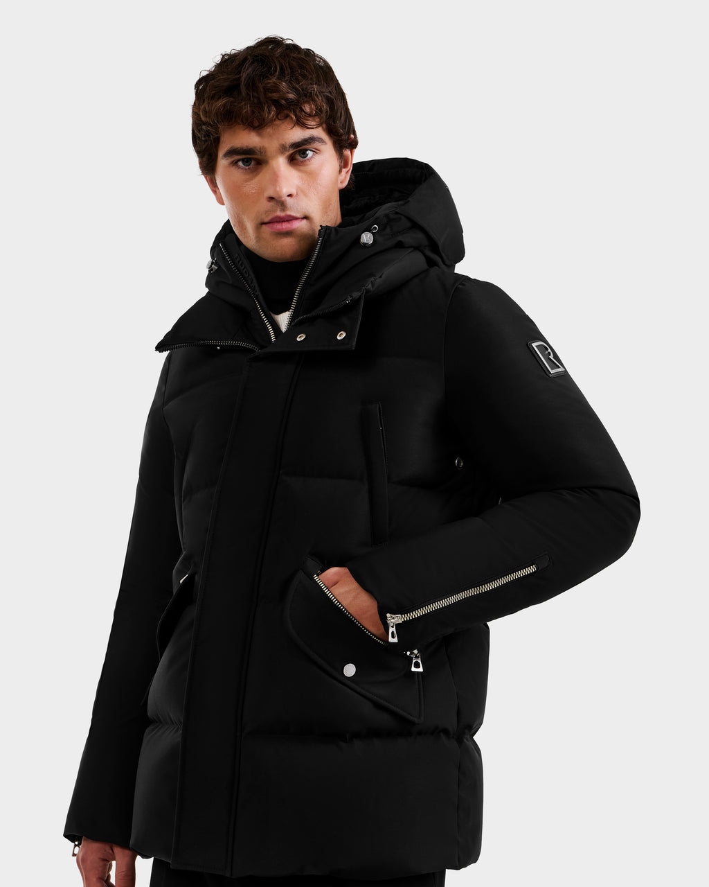 A person wears the Rudsak APOLLO - 6125693 Black parka, featuring silver zippers and an upper sleeve patch, standing slightly sideways with hand in pocket against a plain light background.