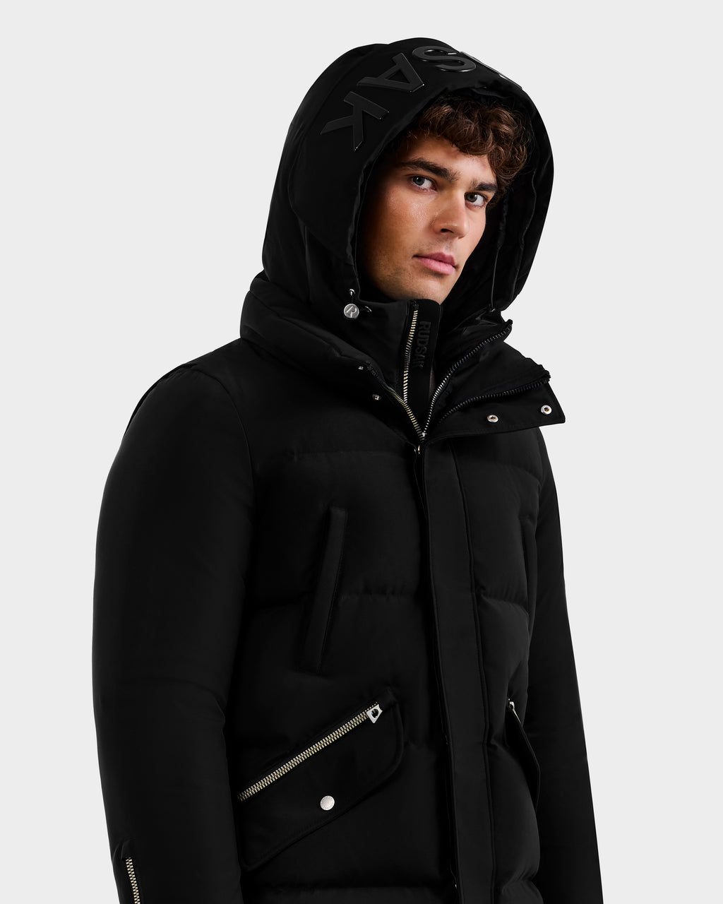 A young man stands against a white background wearing the Rudsak APOLLO - 6125693 Black puffer jacket with a hood, zipper and button details, and large bold lettering on the hood’s edge.