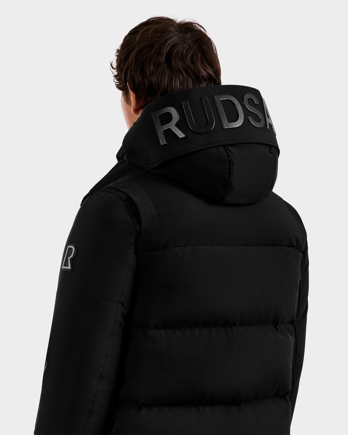 A person faces away, showing the back and side of the APOLLO - 6125693 Black puffer jacket by Rudsak, featuring a large hood with "RUDSAK" in bold black letters.