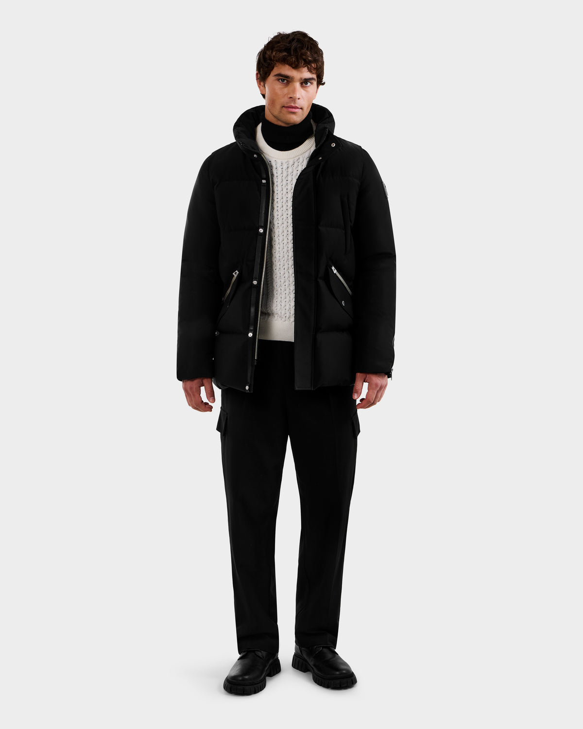 A man faces forward against a light gray background, wearing the Rudsak APOLLO - 6125693 Black puffer jacket over a white knit sweater, paired with black trousers and black shoes.