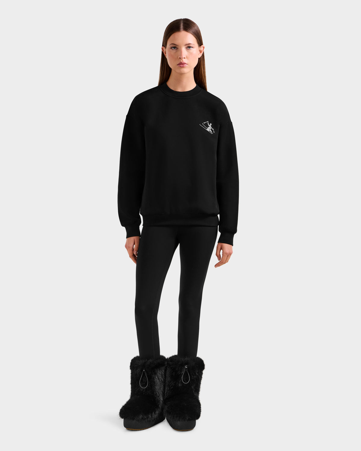 A woman with straight brown hair and a neutral expression wears the Rudsak ARIA - 8525661 Black women's crewneck sweatshirt, featuring a small white logo. She pairs it with black leggings and large black furry boots against a plain background.