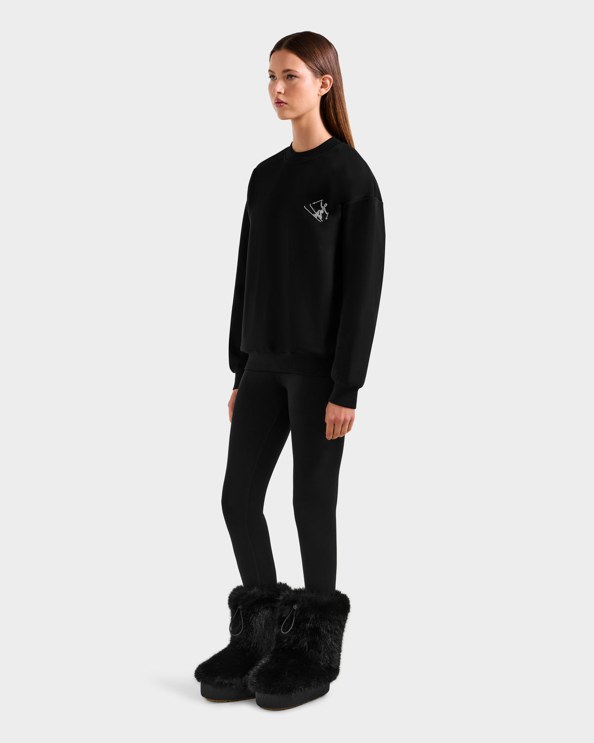 A woman stands against a plain background wearing the Rudsak ARIA - 8525661 Black women’s crewneck sweatshirt, featuring a subtle alpine-inspired white graphic, paired with black leggings and large black furry boots.