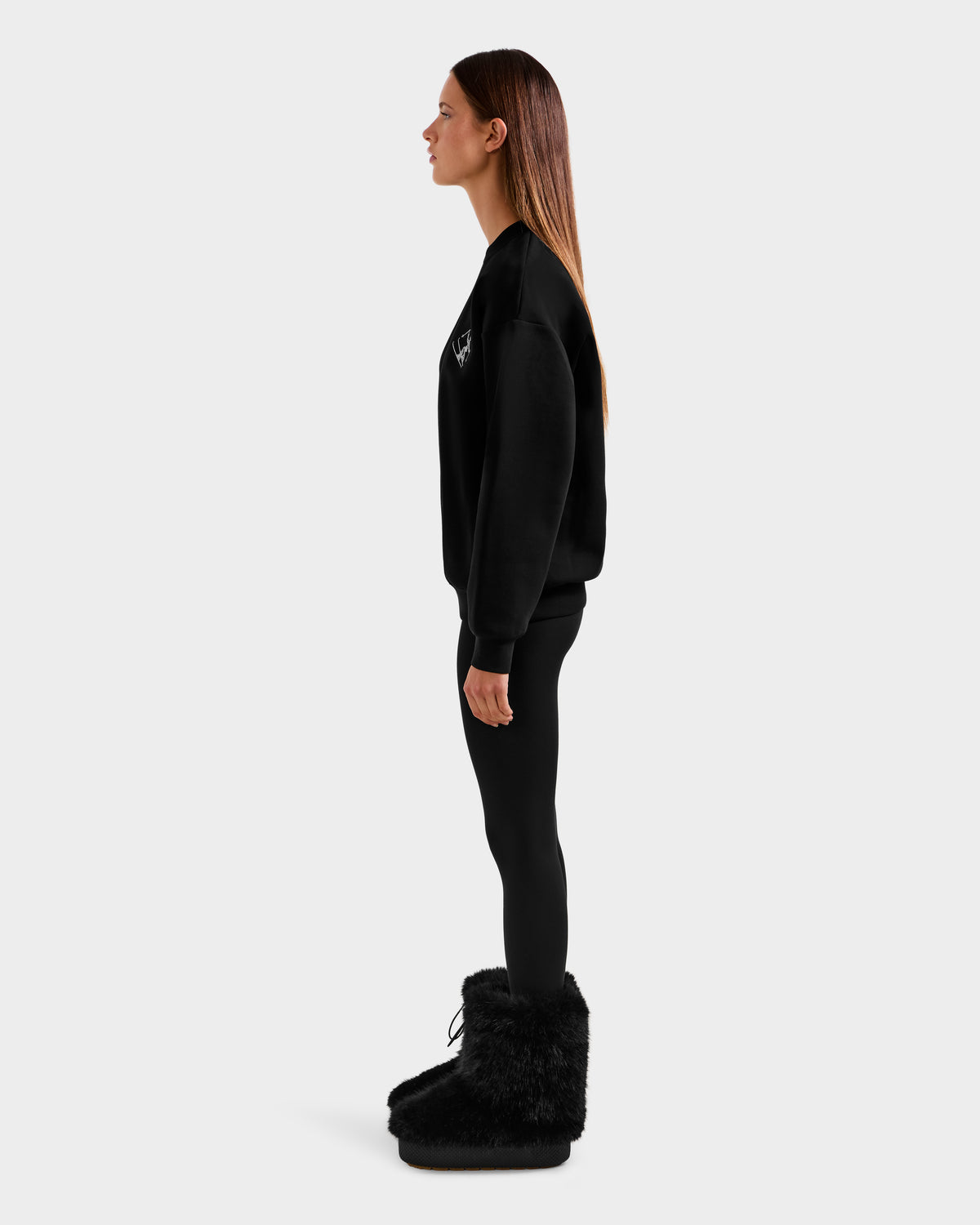 A woman with straight brown hair stands in profile, wearing the ARIA - 8525661 Black sweatshirt by Rudsak, paired with black leggings and large black furry boots against a plain light gray background.