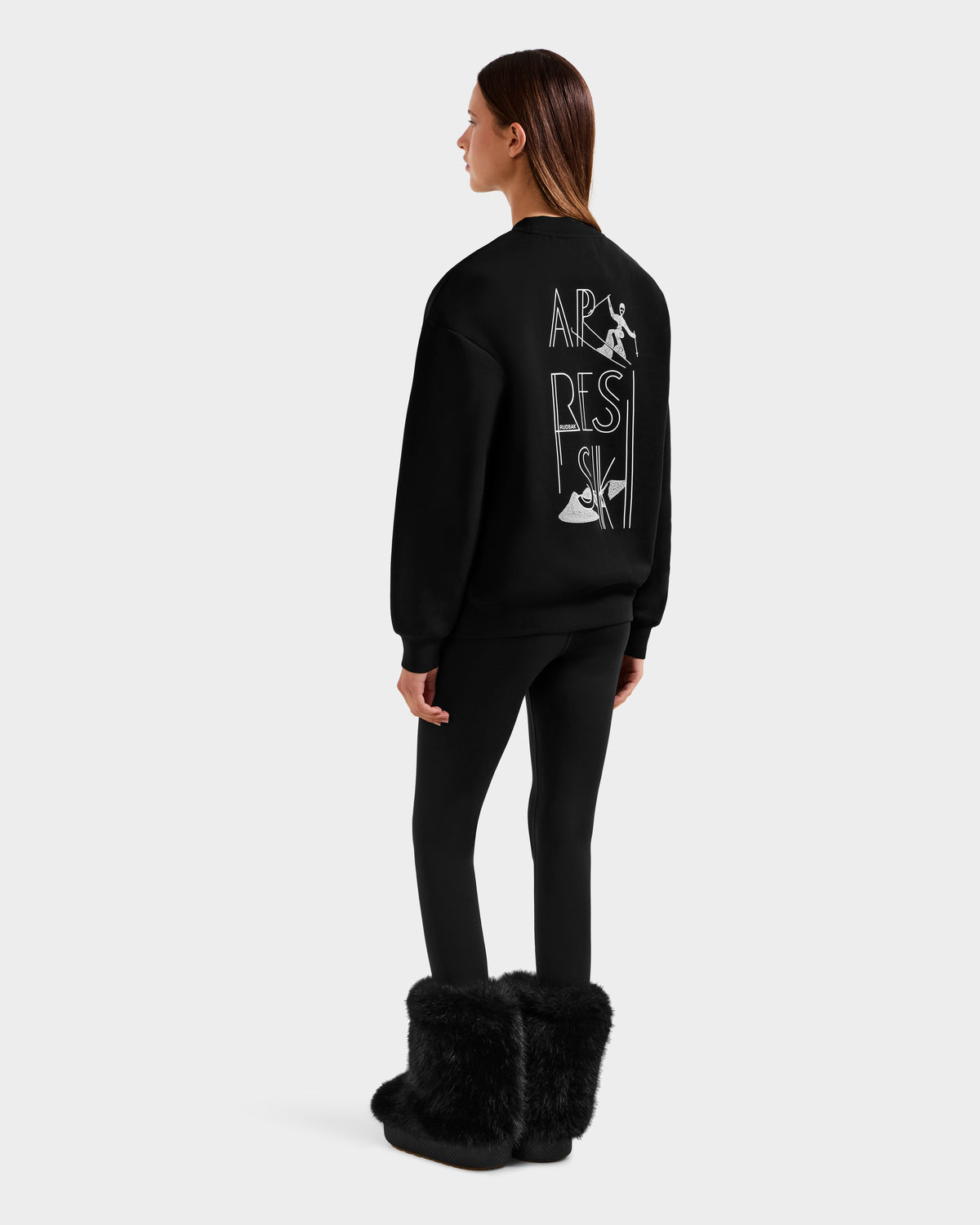 A woman, facing away, wears the Rudsak ARIA - 8525661 Black women’s crewneck sweatshirt with APRES SKI text and graphic on the back, paired with black leggings and furry boots, set against a plain light background.