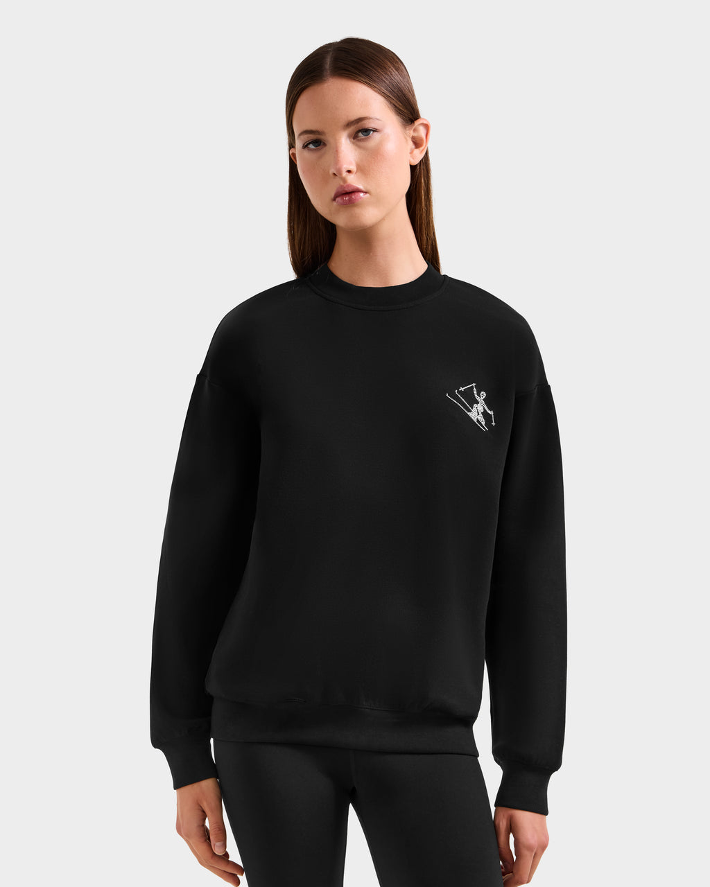 A woman with straight brown hair faces forward, wearing the Rudsak ARIA - 8525661 Black women's crewneck sweatshirt with a small white ski graphic on the left chest and black leggings, set against a plain light gray background.