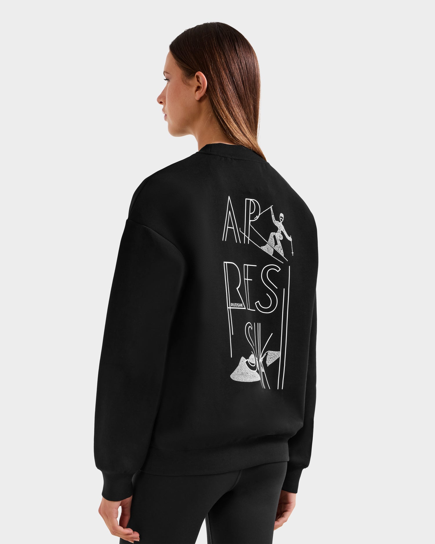 A woman with straight brown hair models the Rudsak ARIA - 8525661 Black women’s crewneck ski sweatshirt, featuring APRES SKI text and alpine-inspired graphics on the back, against a plain light background.
