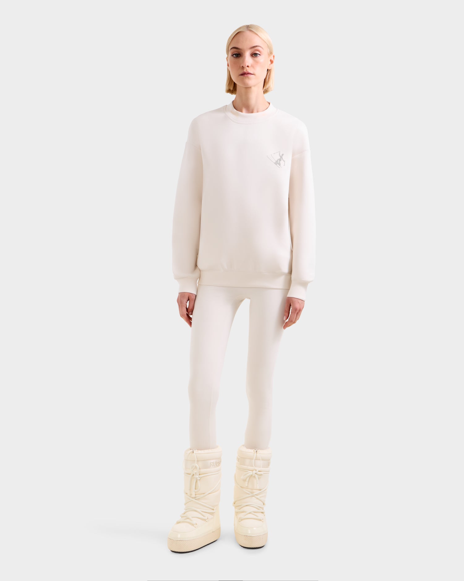 A person faces forward in the Rudsak ARIA - 8525661 Vanilla après-ski pullover with fitted pants and large white snow boots, set against a plain light gray background.