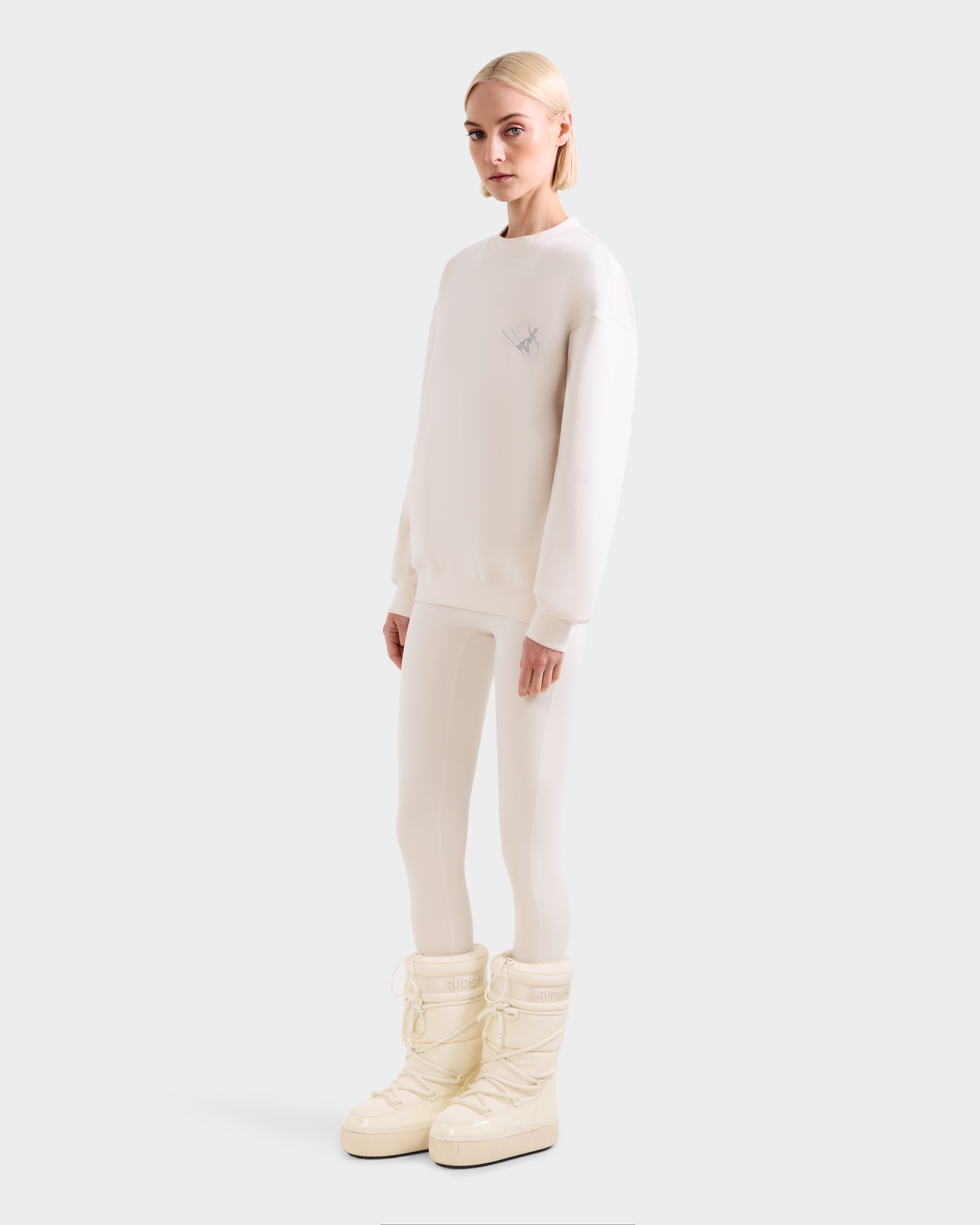 A woman with straight blonde hair stands against a plain background, wearing Rudsak's ARIA - 8525661 Vanilla tall, padded off-white snow boots.