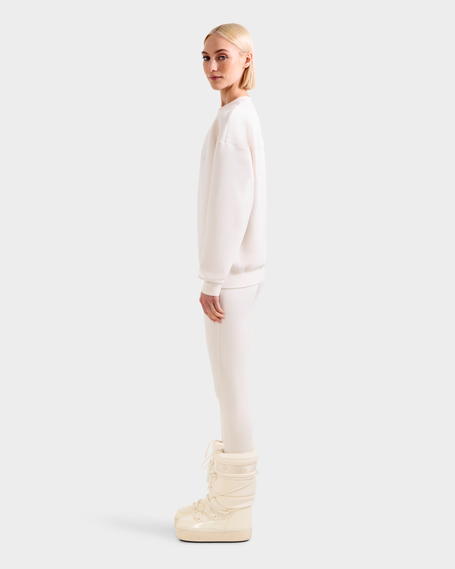 A person with straight blonde hair stands in profile, wearing an oversized women’s crewneck sweatshirt, white leggings, and Rudsak ARIA - 8525661 Vanilla cream puffy snow boots against a plain light gray background.