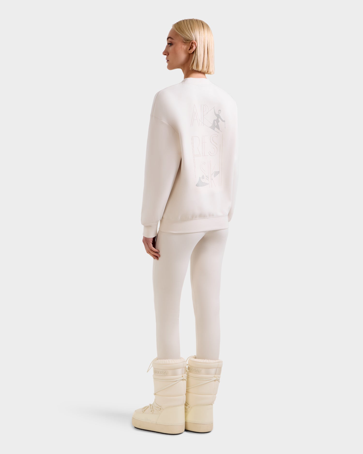 A person with straight blonde hair faces away, dressed in all white, wearing the Rudsak ARIA - 8525661 Vanilla women's crewneck sweatshirt featuring subtle graphic text and images on the back, paired with matching leggings and cream winter boots.