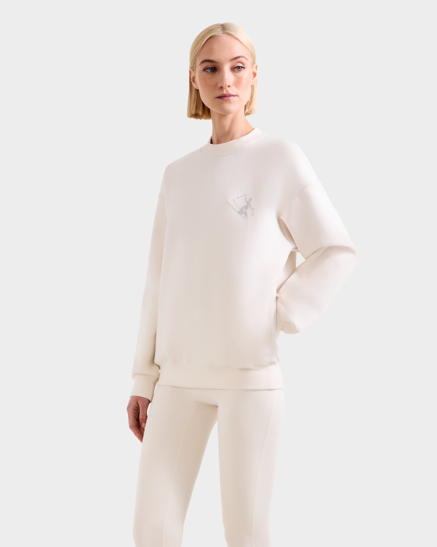 A blonde woman stands against a plain light background wearing the ARIA - 8525661 Vanilla women’s crewneck sweatshirt and matching pants by Rudsak, with one hand on her hip.