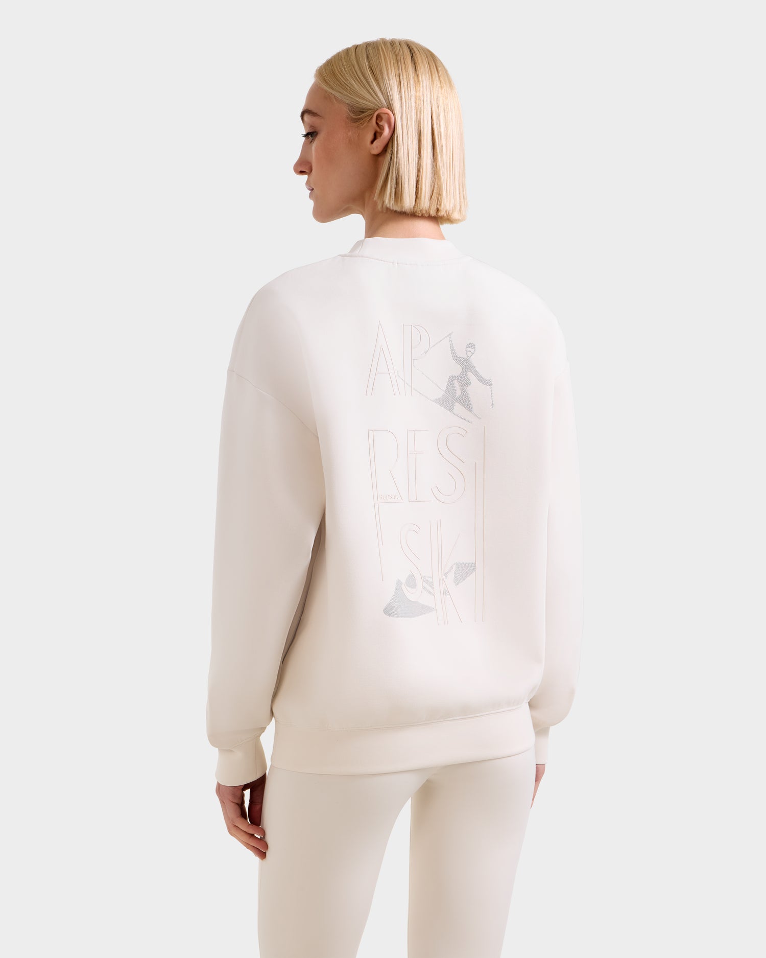 A woman with straight blonde hair wears the Rudsak ARIA - 8525661 Vanilla women’s crewneck sweatshirt, shown from the back. This off-white top features subtle ski-themed graphics and "APRÈS SKI" text, paired with leggings.
