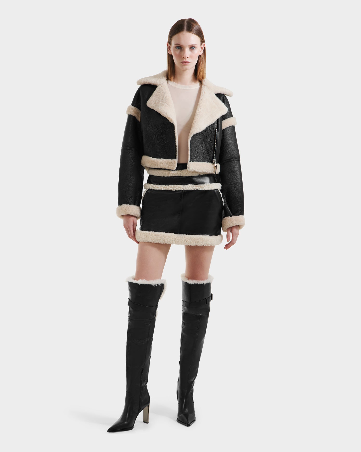 A woman faces forward against a white background, wearing the Rudsak ARIANNA - 8125504 Black minimalist cropped shearling jacket with white lining, a matching mini skirt, and knee-high black heeled boots.