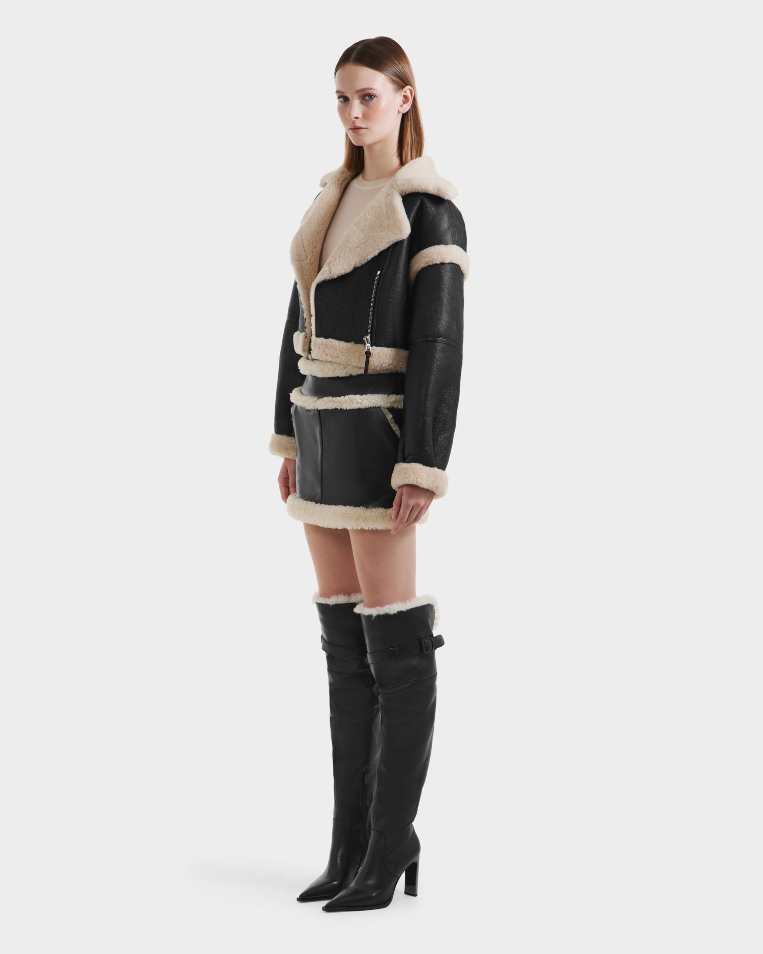 A woman faces left against a plain white background, wearing Rudsak ARIANNA - 8125504 Black knee-high heeled boots, styled with a cropped shearling jacket and a mini skirt featuring beige shearling and leather trim.