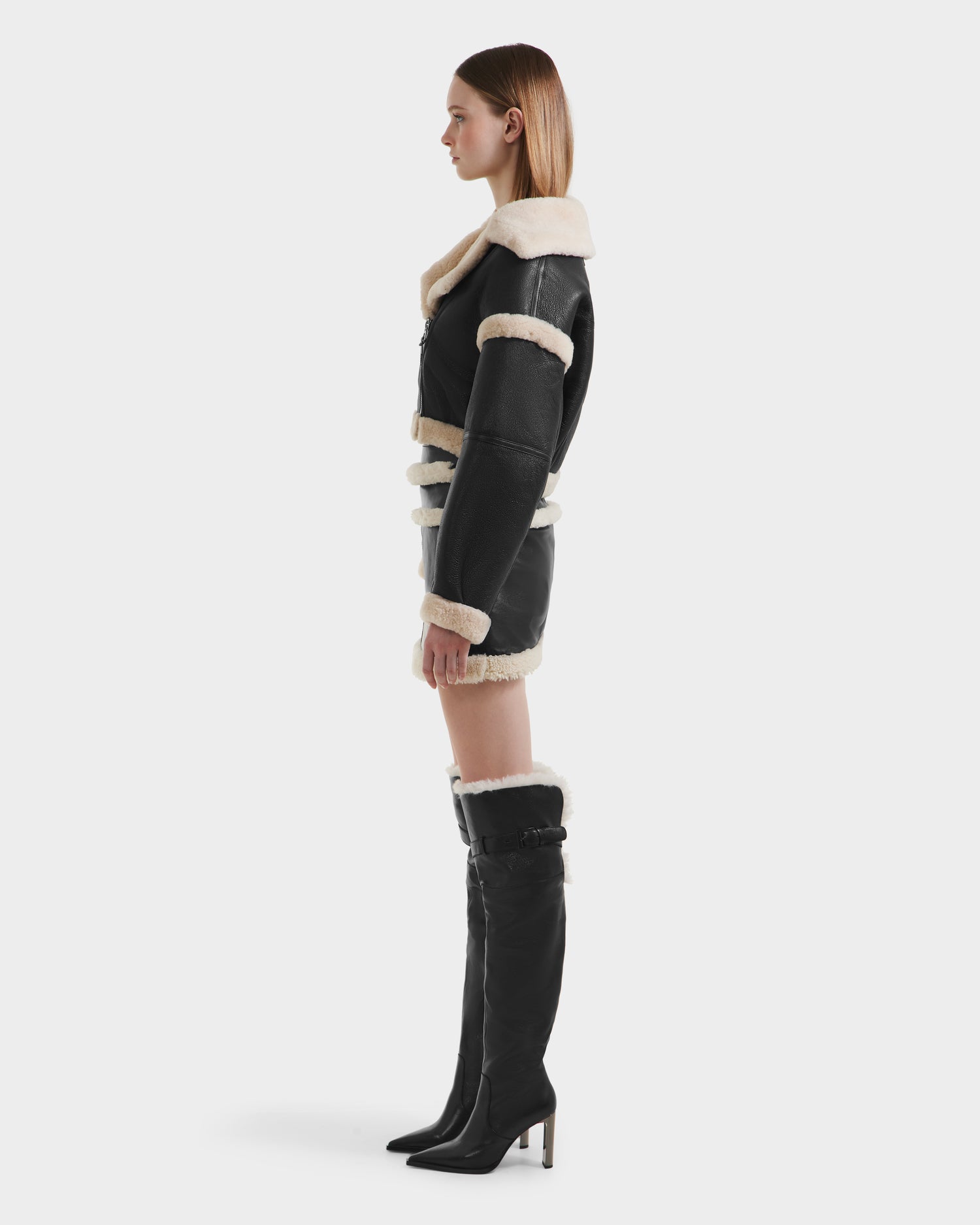 A woman stands in profile against a white background wearing the Rudsak ARIANNA - 8125504 Black minimalist cropped shearling jacket with white leather trim, paired with a matching mini skirt and knee-high black boots featuring shearling accents.