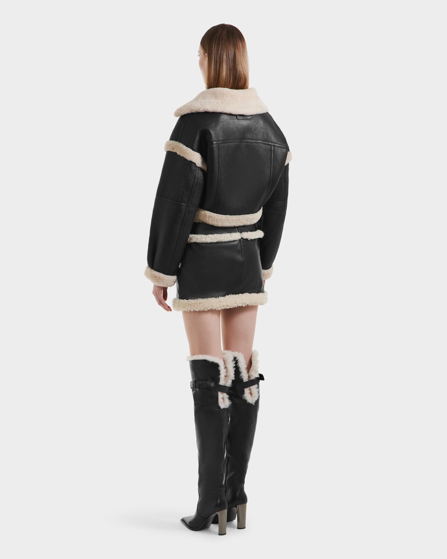A person stands facing away, wearing the Rudsak ARIANNA - 8125504 Black tall boots with fur lining, paired with a cropped shearling jacket and matching mini skirt. The minimalist background is plain white.