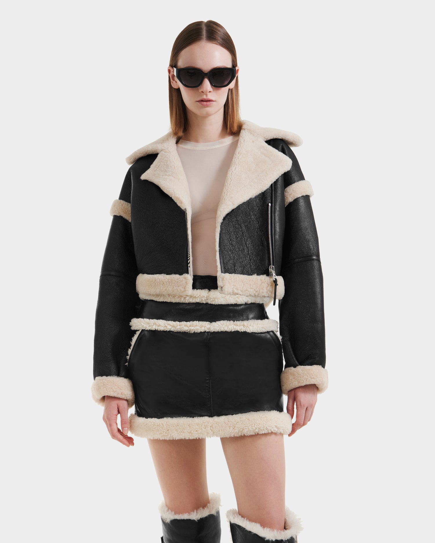 A woman in Rudsak’s ARIANNA - 8125504 Black cropped shearling jacket and matching mini skirt with cream trim, beige top, knee-high boots, and sunglasses poses in a modern minimalist style against a white background.