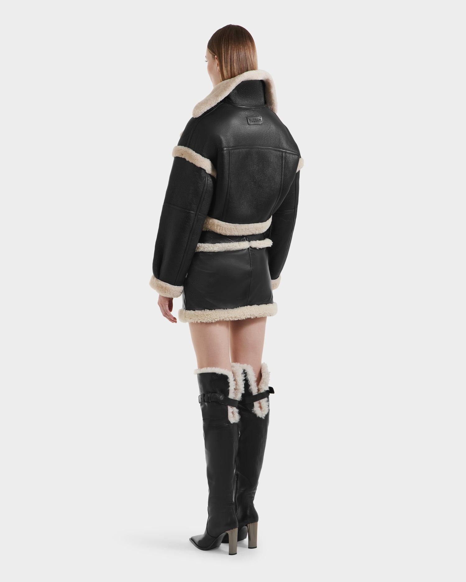 A woman faces away, wearing the Rudsak ARIANNA - 8125504 Black cropped shearling jacket with cream trim, a coordinating black mini skirt, and knee-high black boots featuring cream shearling lining.
