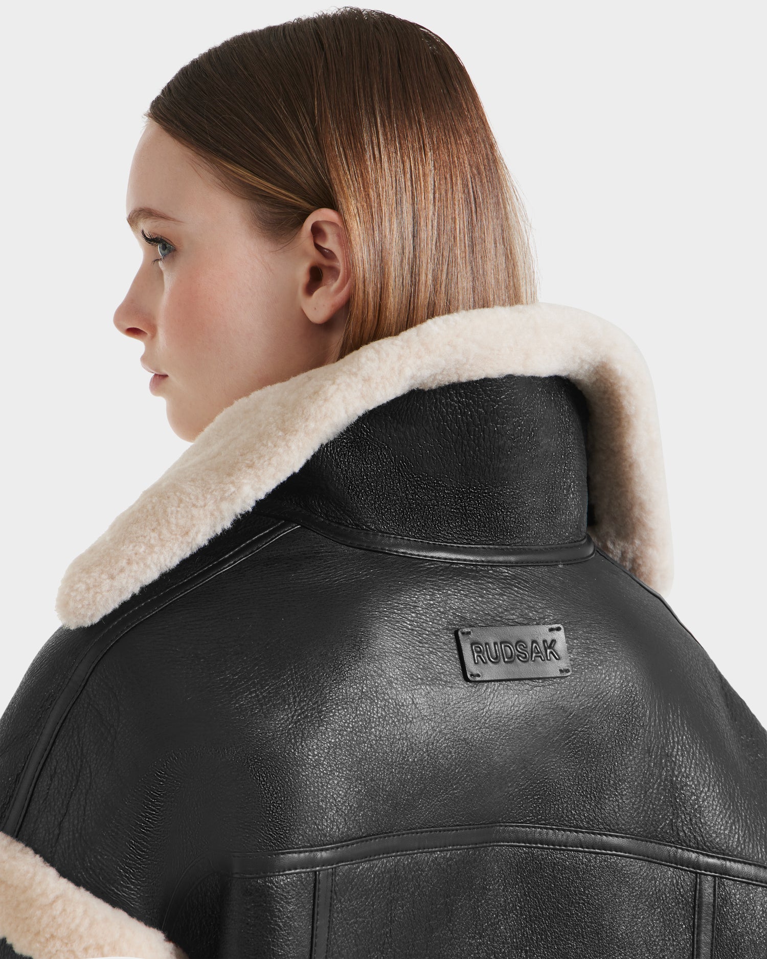 A woman with straight brown hair looks to the side, wearing the Rudsak ARIANNA - 8125504 Black cropped shearling jacket featuring a cream collar, leather trim, and a RUDSAK label on the back.