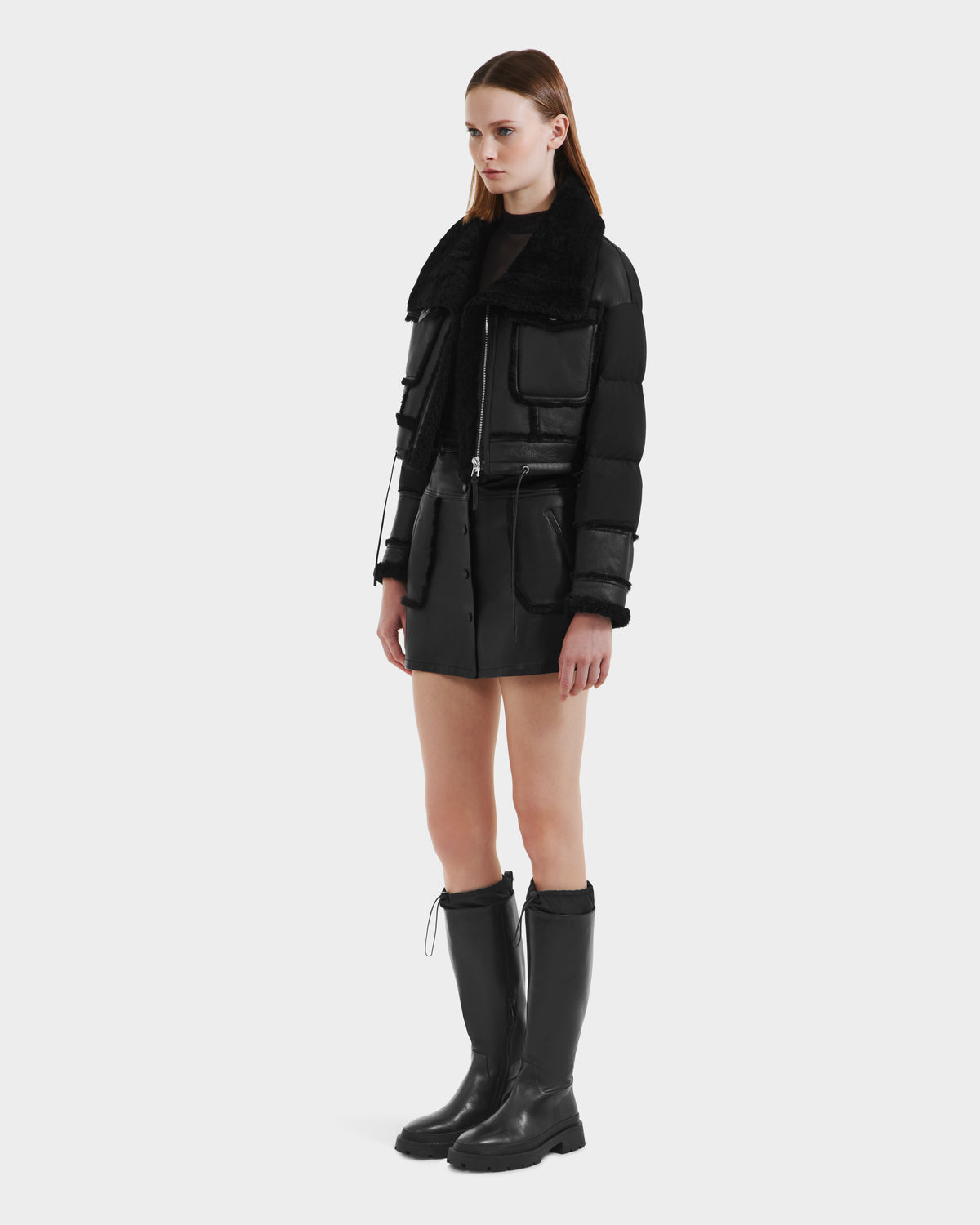 A woman with straight, shoulder-length hair and a neutral expression wears the Rudsak ARIELLA - 8125505 Black women’s cropped shearling puffer with large pockets, styled with a black mini skirt and knee-high boots against a plain background.