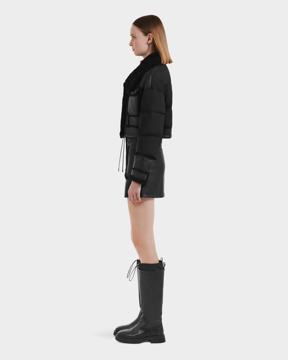 A woman in profile models the Rudsak ARIELLA - 8125505 Black down-filled jacket with a black mini skirt and knee-high drawstring boots, showcasing effortless cold-weather style against a plain light background.