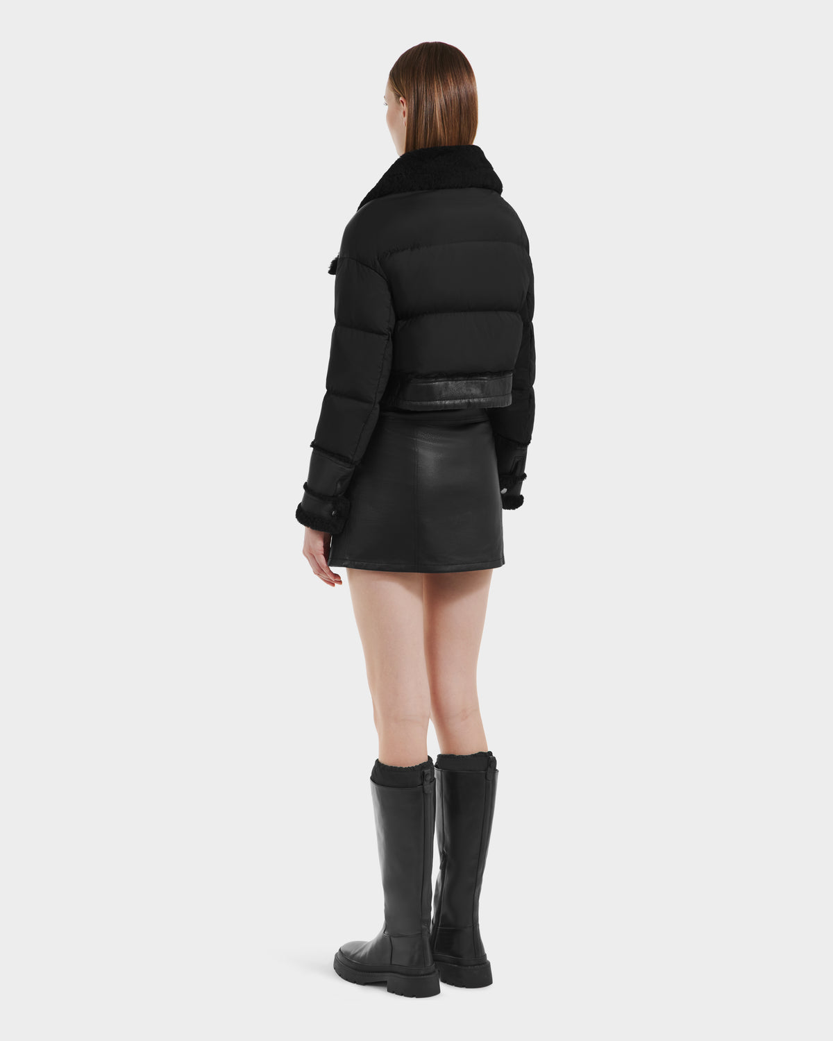 A woman faces away, wearing the ARIELLA - 8125505 Black down-filled jacket by Rudsak with a faux fur collar, paired with a black leather mini skirt and knee-high boots. Her straight brown hair completes her stylish look against a plain white backdrop.