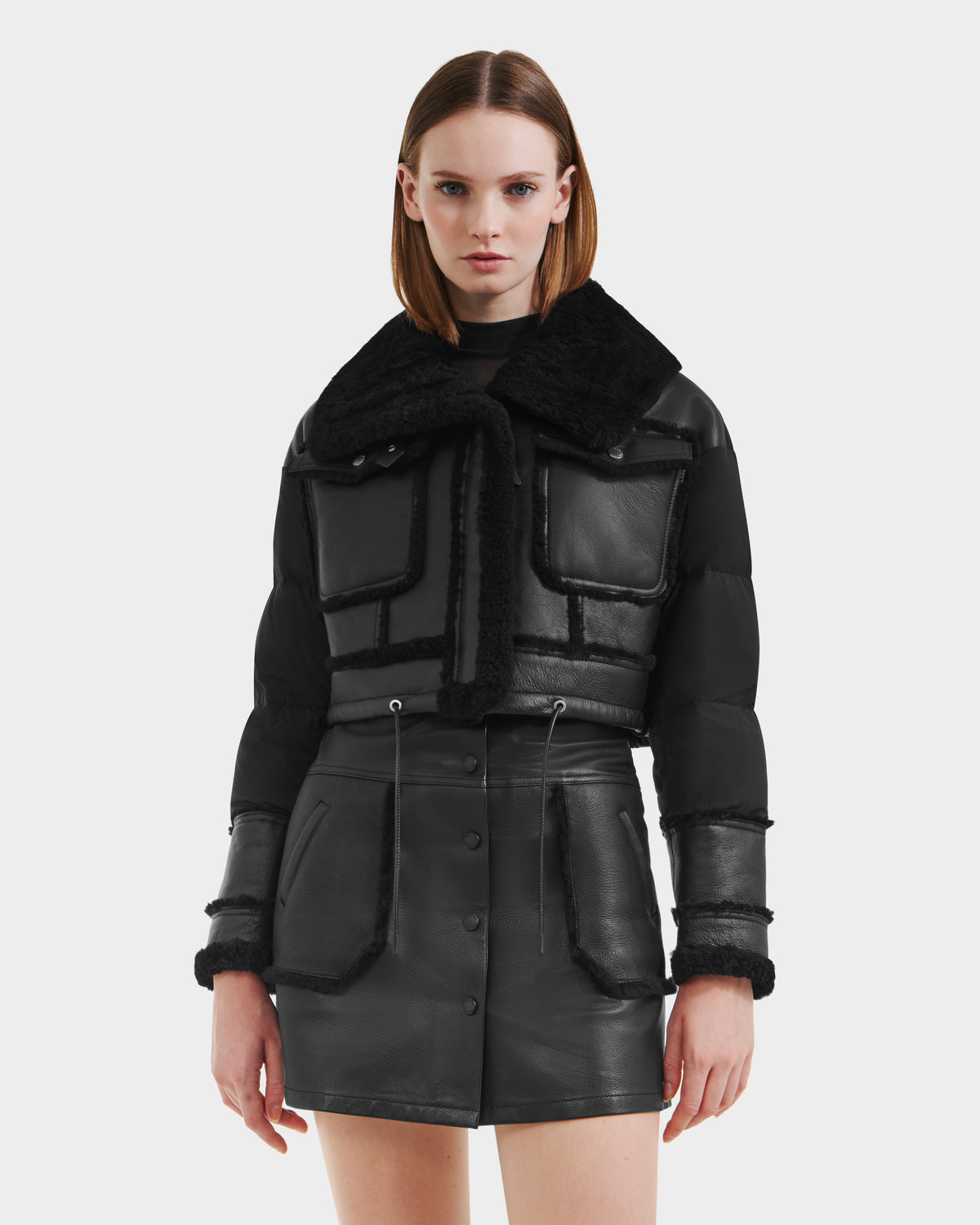 A woman models the Rudsak ARIELLA - 8125505 Black jacket with shearling trim and a matching black leather skirt, standing against a white background to showcase chic cold-weather style.