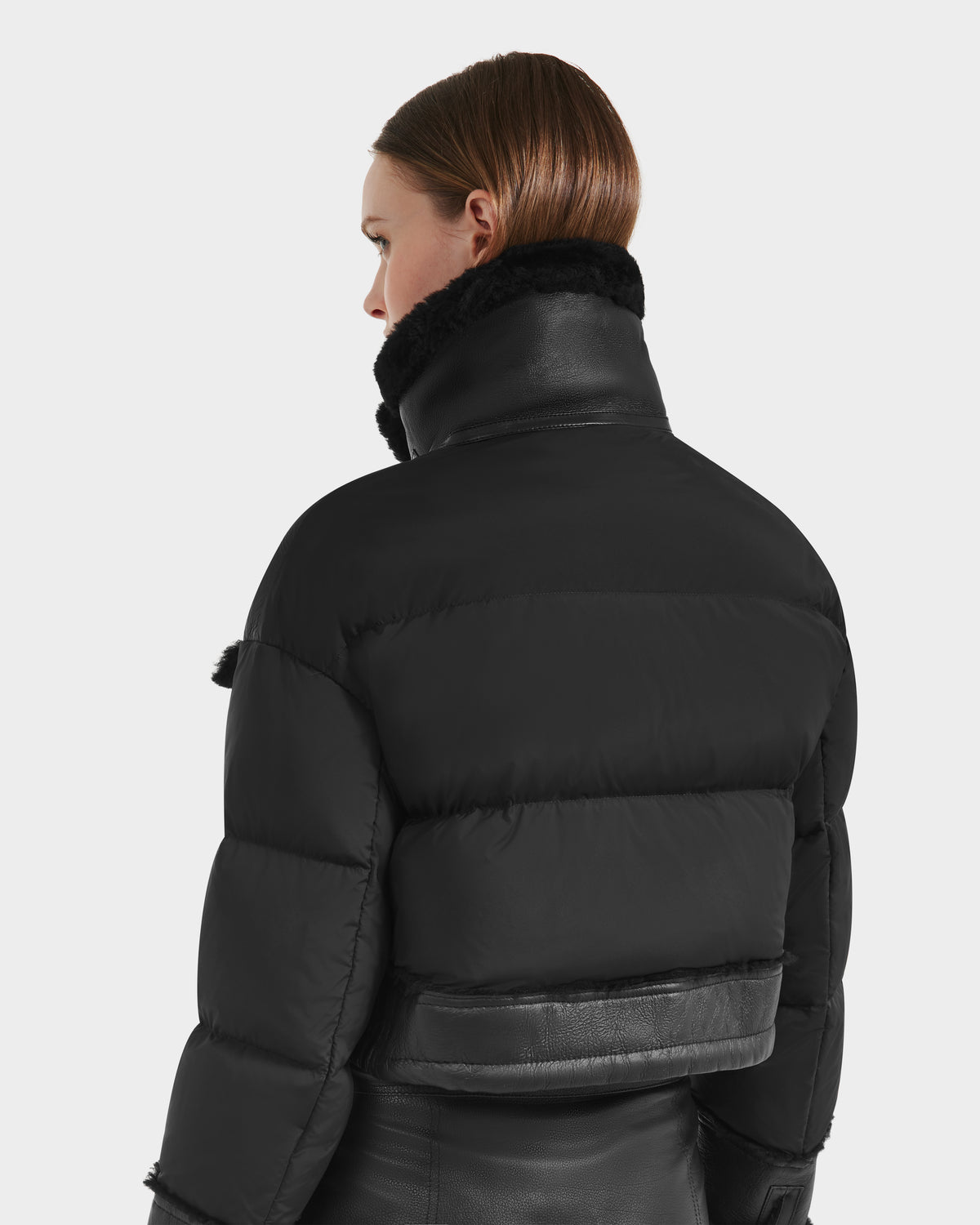 A person with straight brown hair models the Rudsak ARIELLA - 8125505 Black women’s cropped shearling puffer, featuring a high collar and leather accents, against a plain light background—perfect for cold-weather styling.
