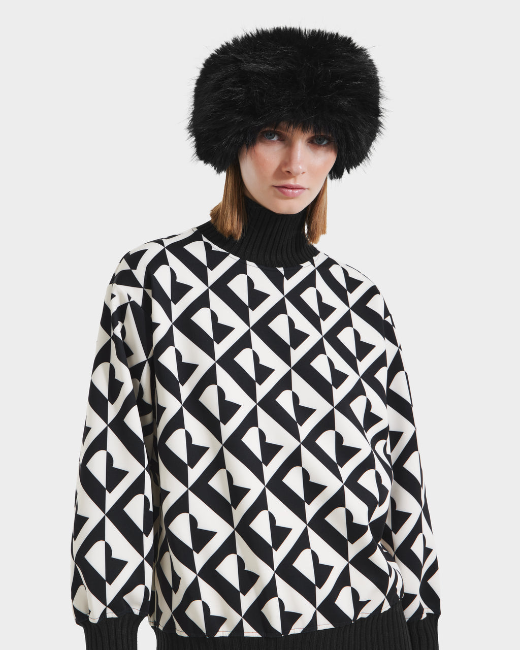 A woman models the Rudsak ARLETTE - 8425544 Black faux fur hat, paired with a black-and-white geometric heart sweater over a black turtleneck, standing against a plain white background.