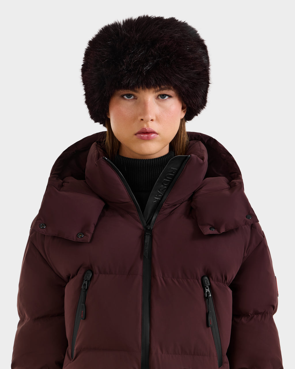 Wearing the Rudsak ARLETTE - 8425544 Bordeaux puffer jacket and a dark brown fur hat, a person in a black turtleneck stands against a plain light background.